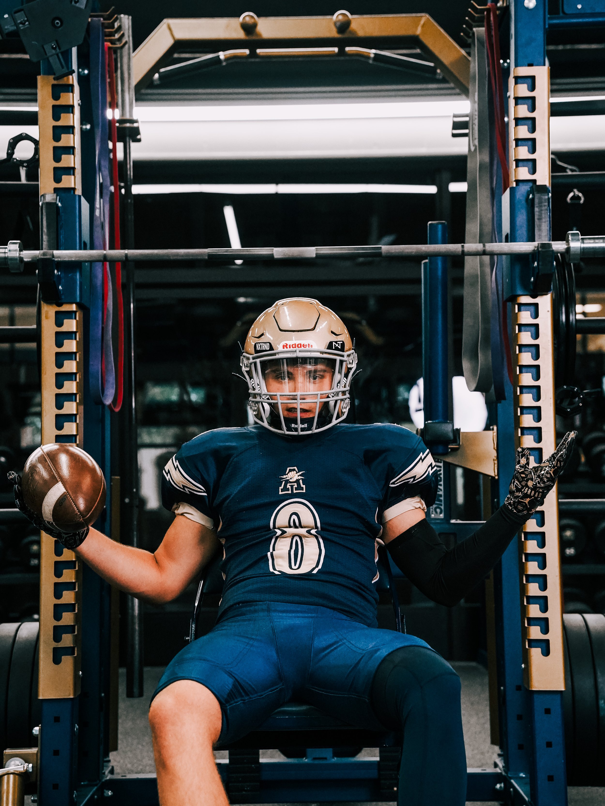A football player in uniform and helmet sitting in a weightlifting cage, holding a football in one hand and flexing his muscles.