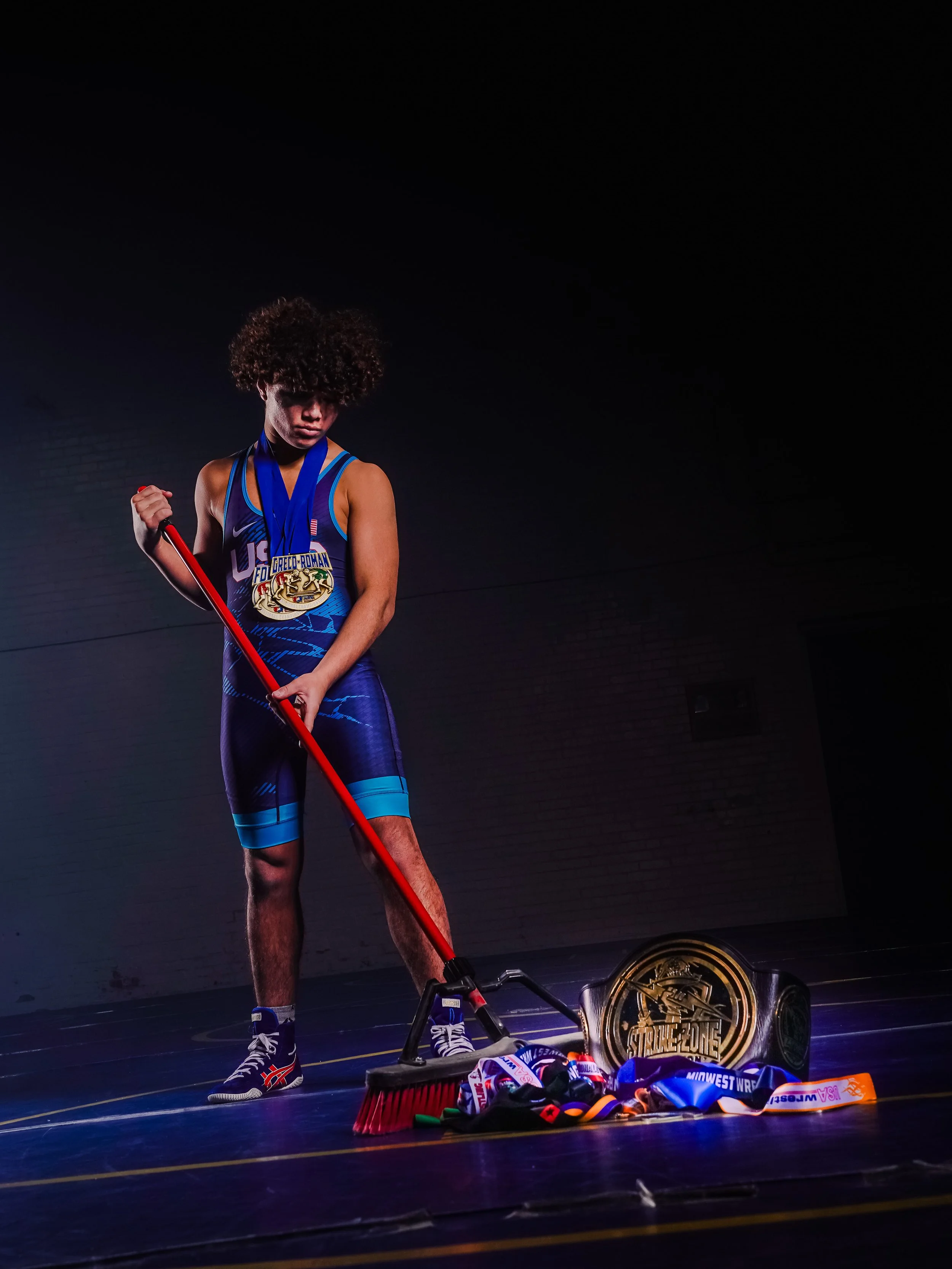 A young male athlete in a blue sports uniform and sneakers stands on a wrestling mat, holding a red broomstick, surrounded by medals and a championship belt, with a dark background.