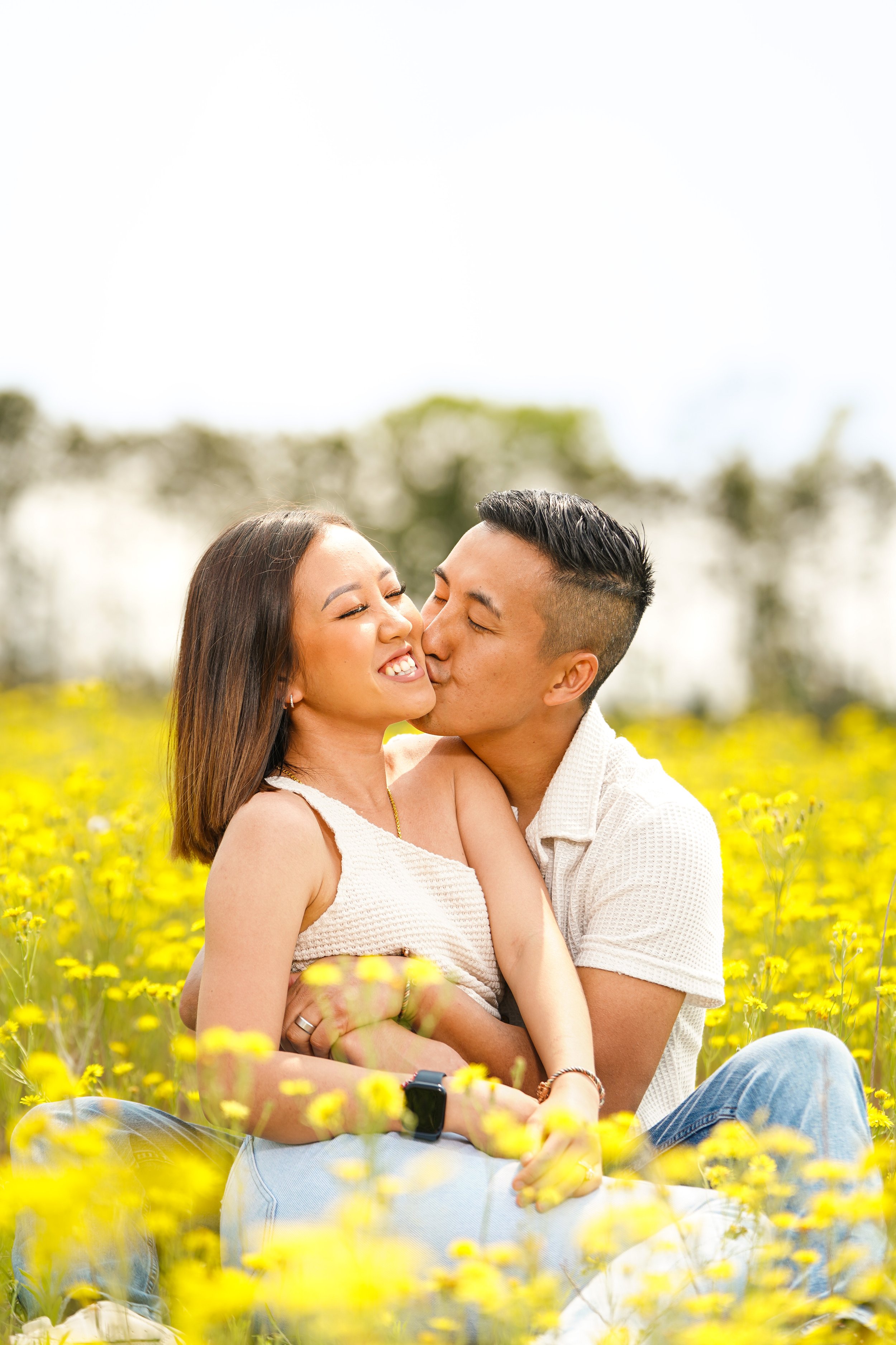 A happy couple sitting in a field of yellow flowers, kissing and smiling.