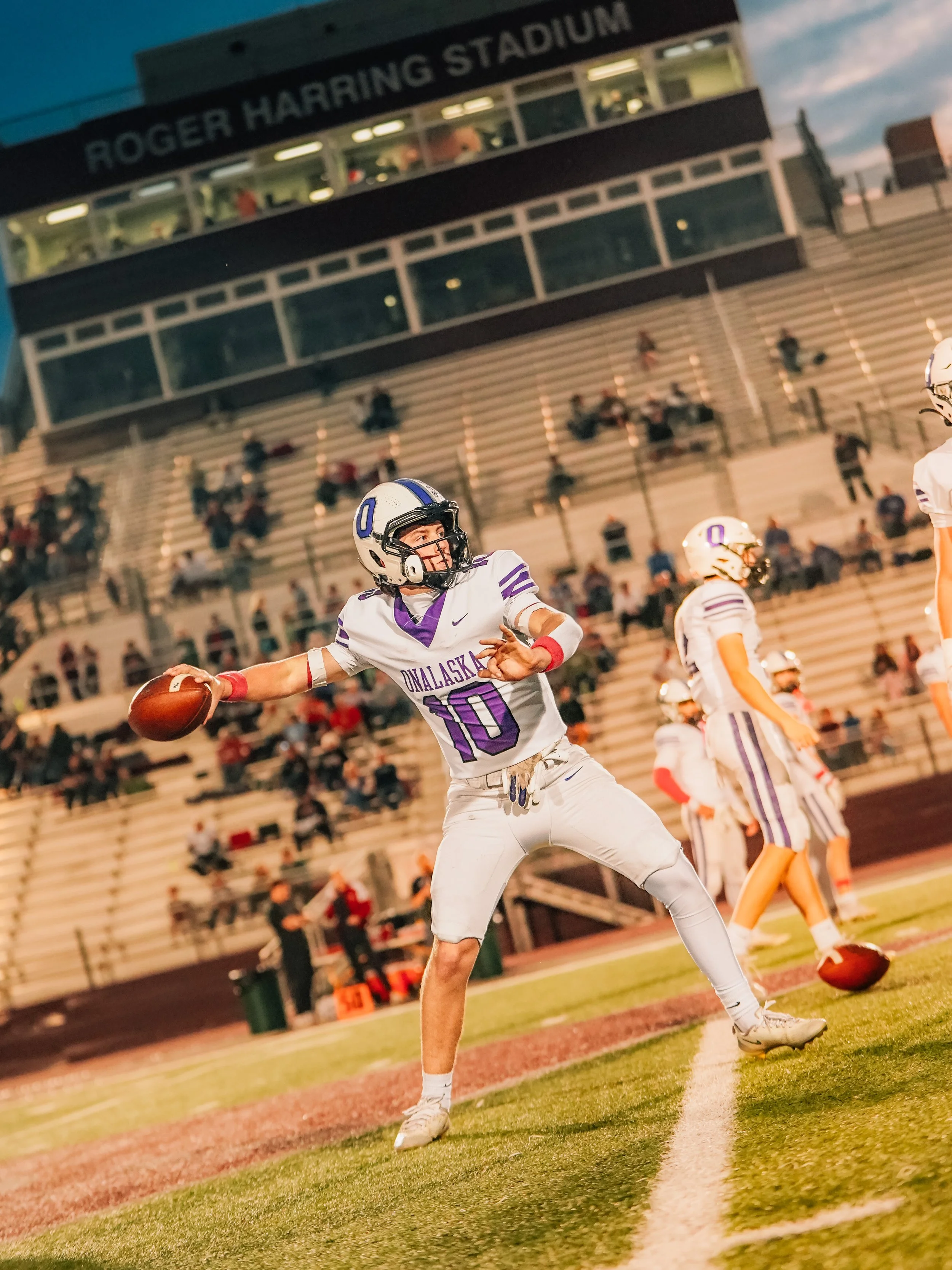 A football player in a white and purple uniform with the number 10 on it, running with the football on a football field during a game at Roger Harring Stadium at night.