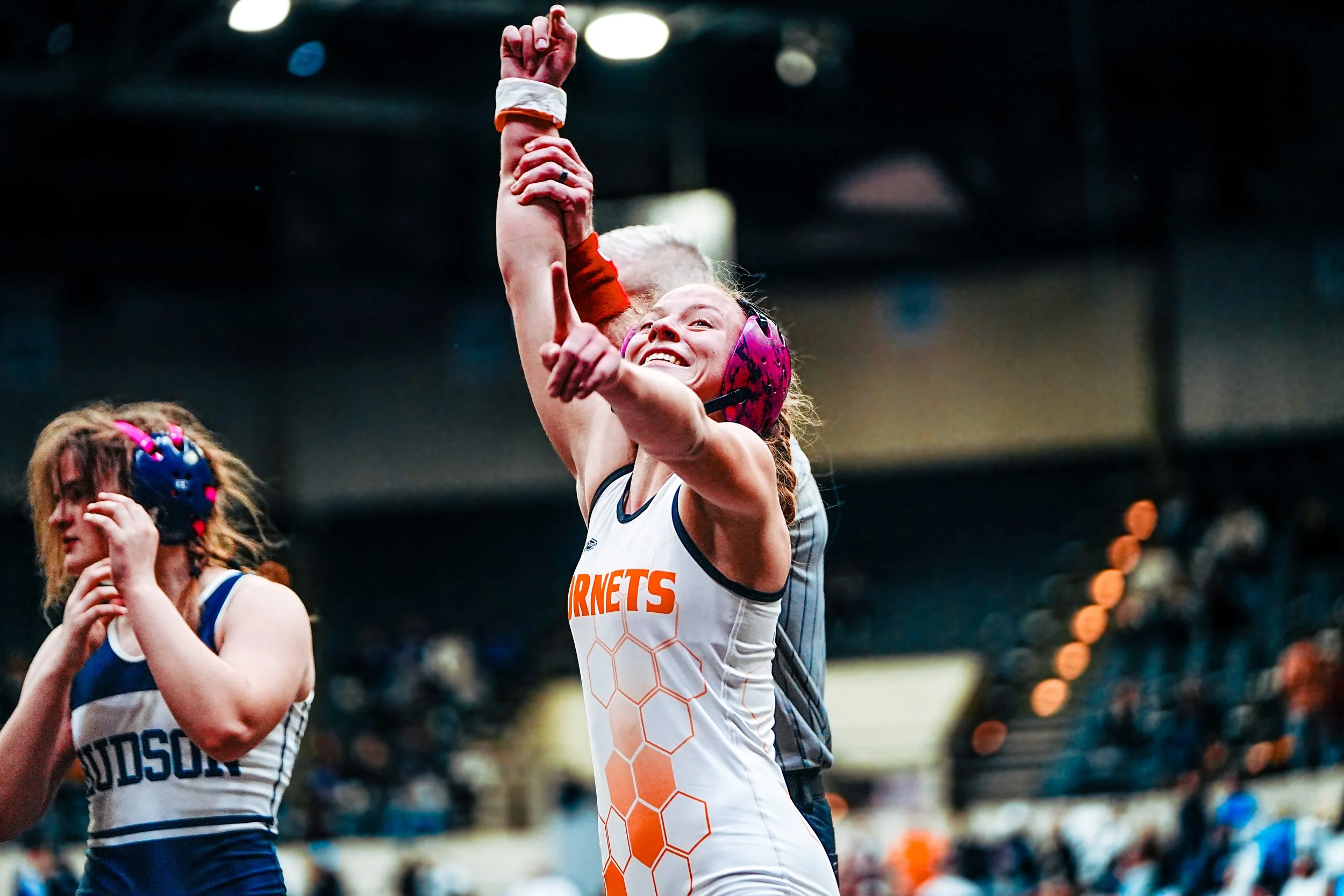 A female roller derby player wearing a white jersey with orange and black hexagon patterns, pink helmet, and black wrist guards, smiling and raising her hand in a sporting arena.