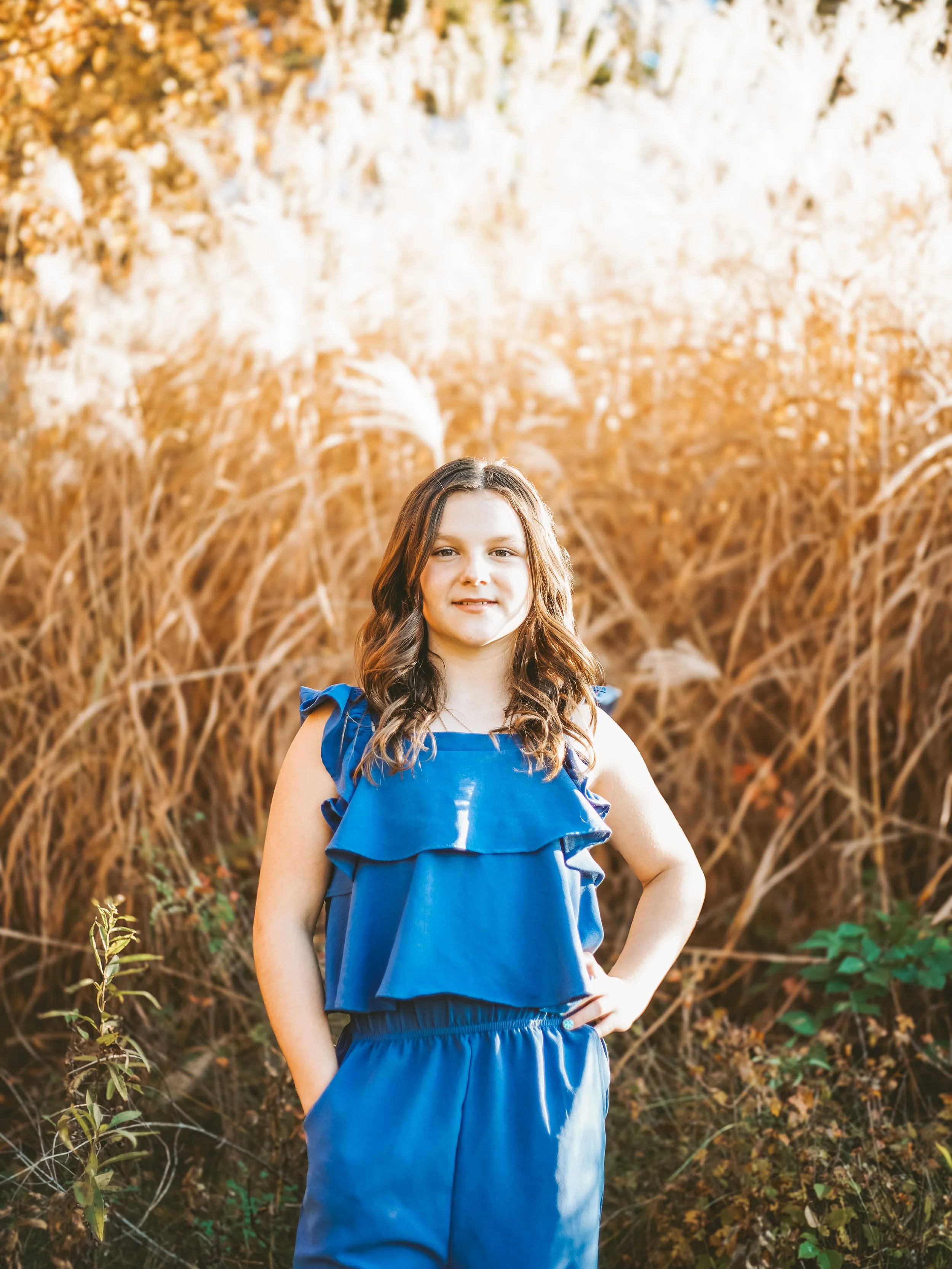 A girl standing outdoors in front of tall, brownish grass with a few green plants, wearing a blue dress, with long brown hair and smiling.