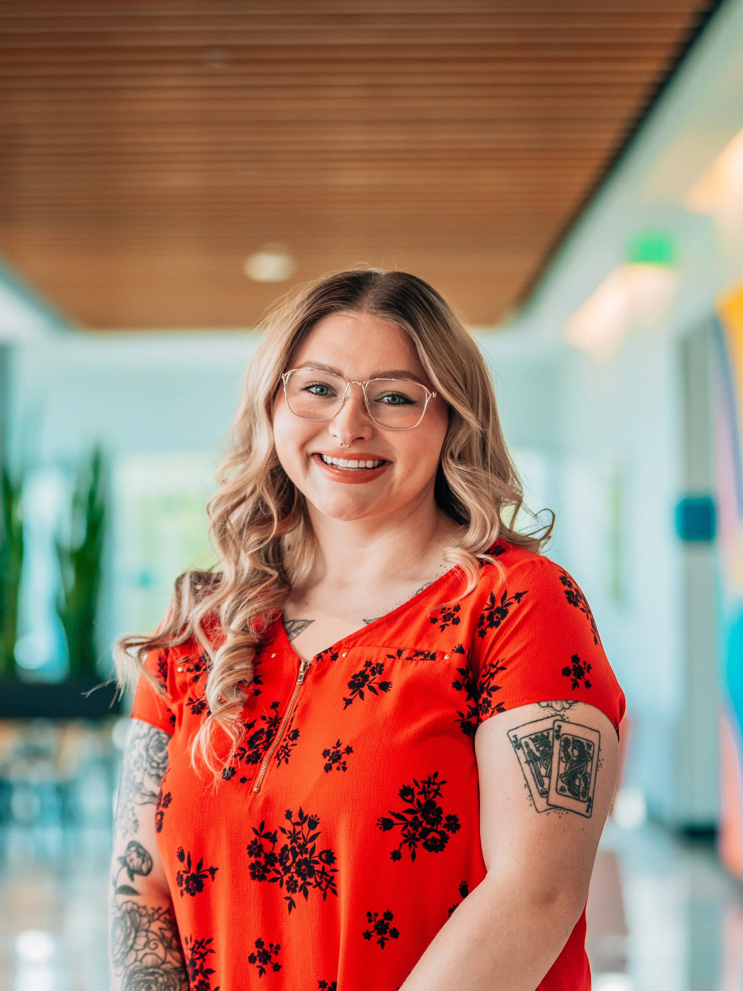 A smiling woman with glasses, tattoos on her arms, and a septum piercing, wearing a red dress with black floral patterns, standing in an indoor setting with a wooden ceiling and blurred background.