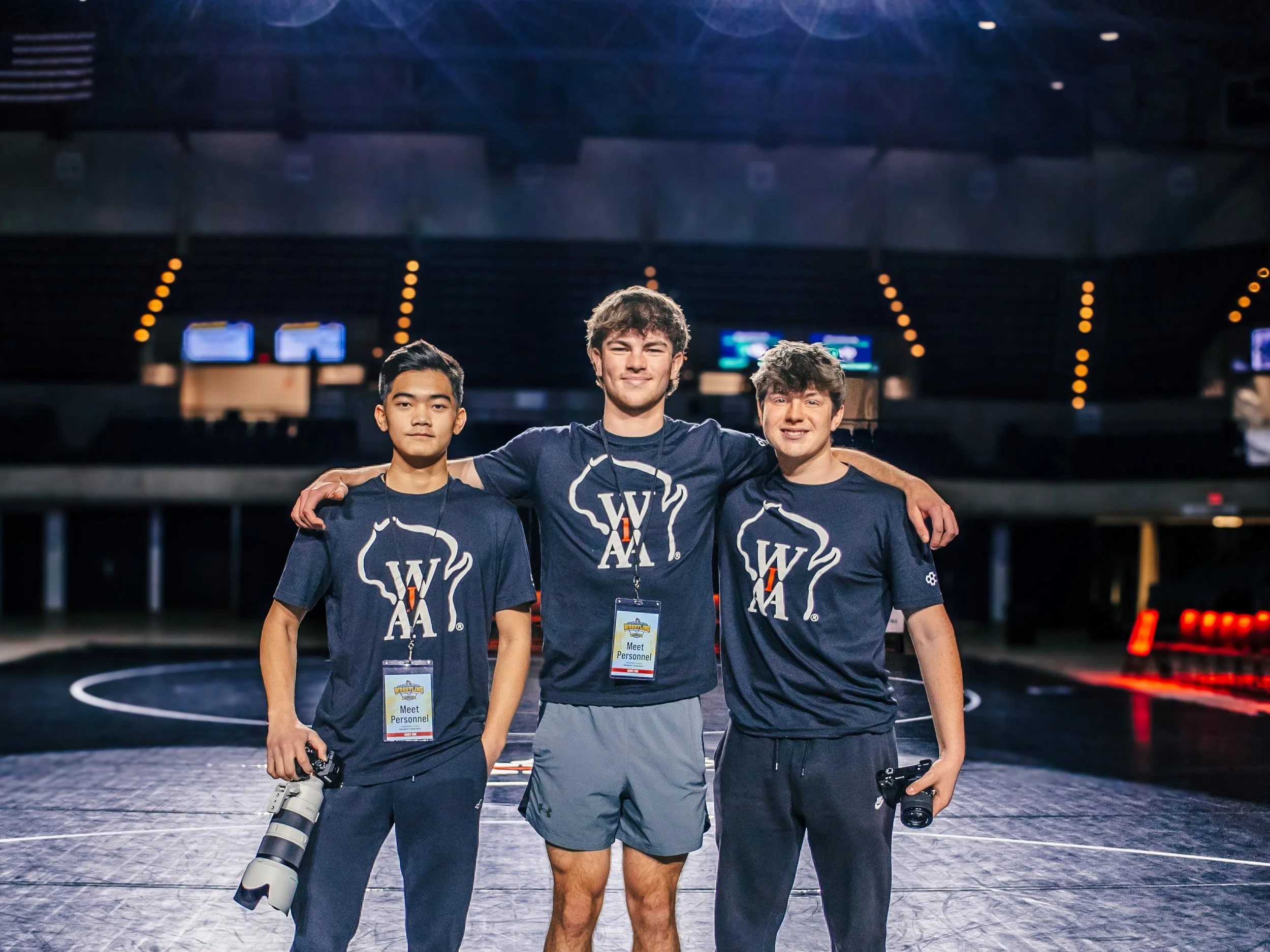 Three young men standing together in a sports arena, wearing matching dark blue t-shirts with a white logo and credentials around their necks, smiling at the camera, one holding a camera with a large lens.