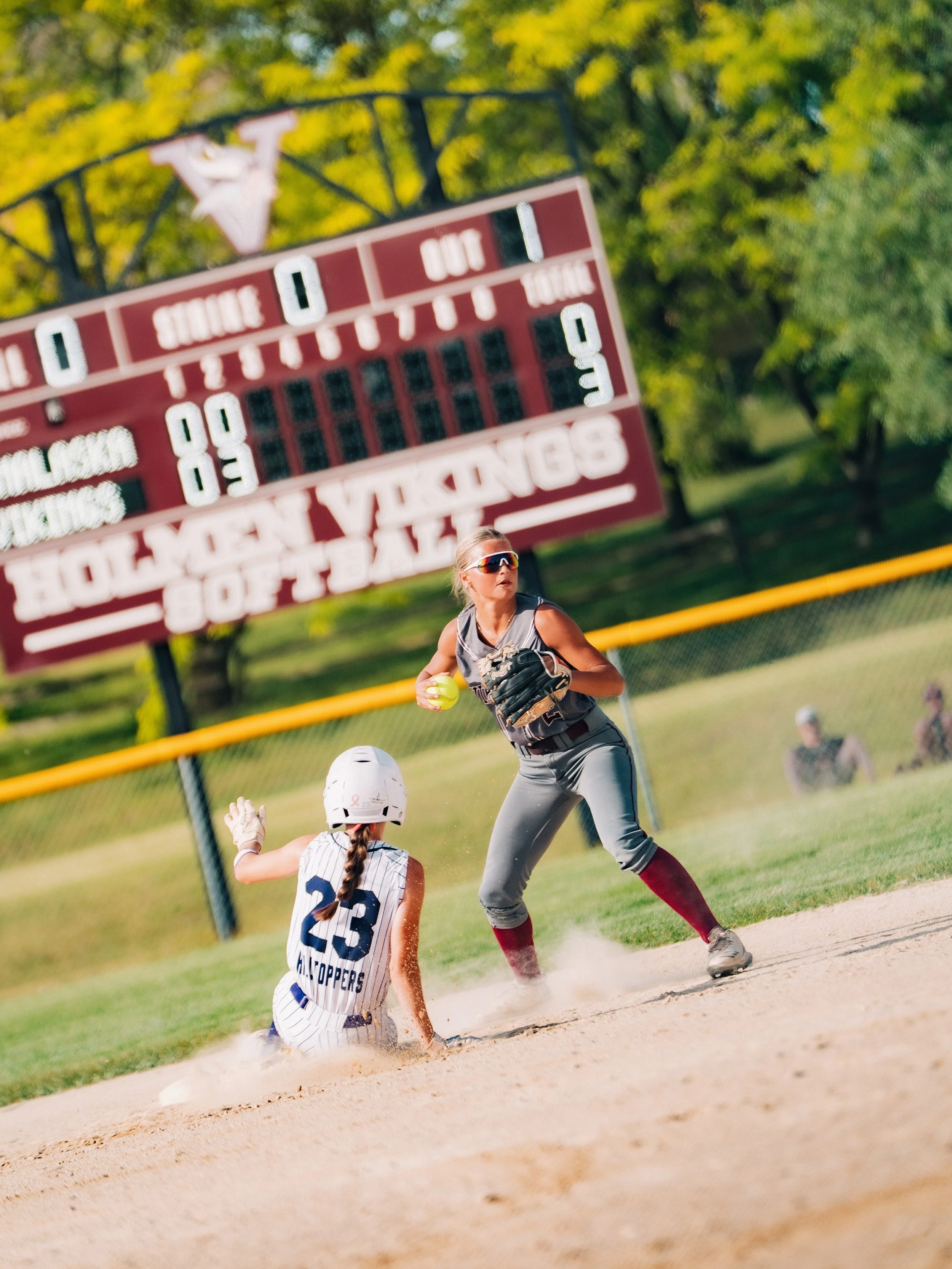 A softball player in gray and red uniform sliding into home plate while a player in a striped uniform and helmet attempts to tag her out in a sunny park with trees in the background.