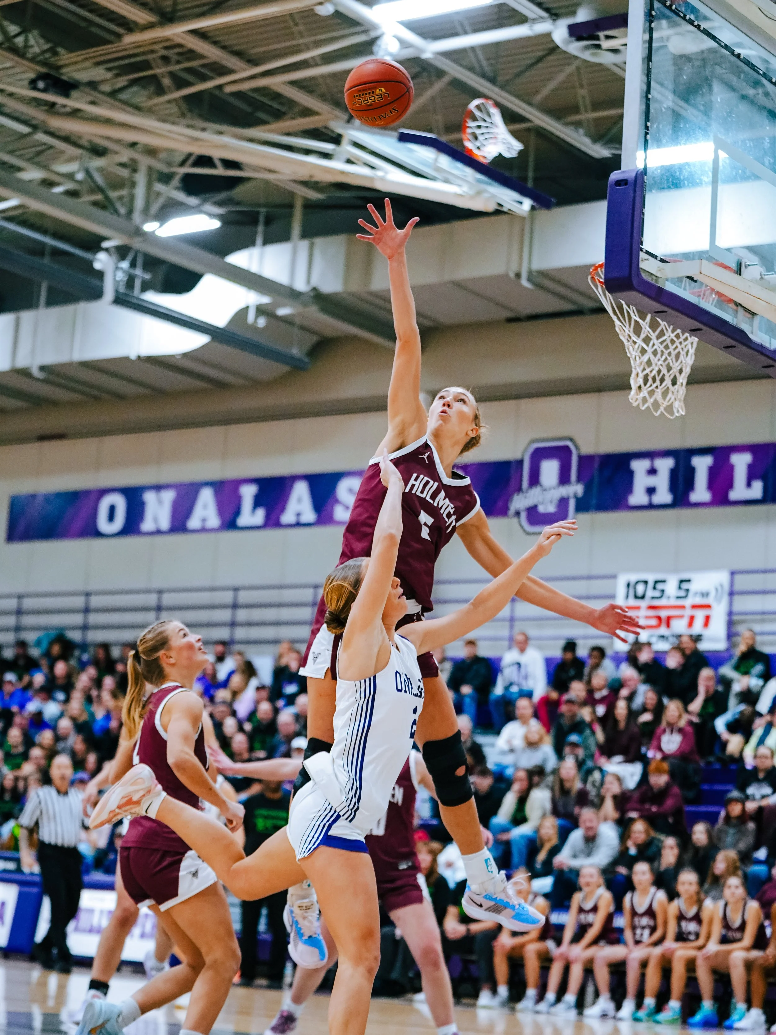 A woman basketball player in a maroon jersey is jumping to shoot or block in a game, with other players and spectators in the background.
