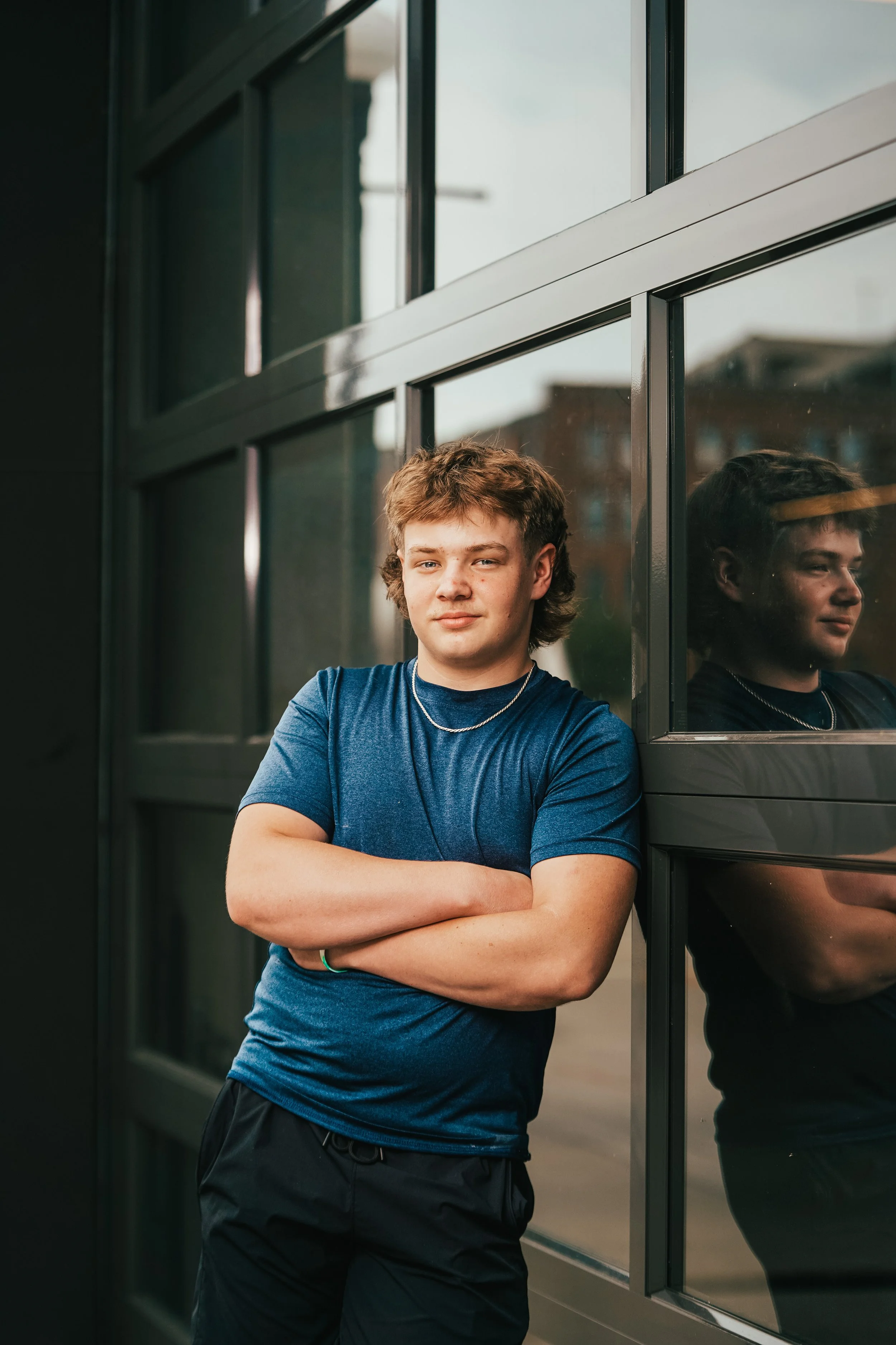A young man with light brown, curly hair and blue eyes stands outside next to a building with reflective glass windows. He is wearing a blue T-shirt, a necklace, and black pants, and has his arms crossed. His reflection is visible in the glass window