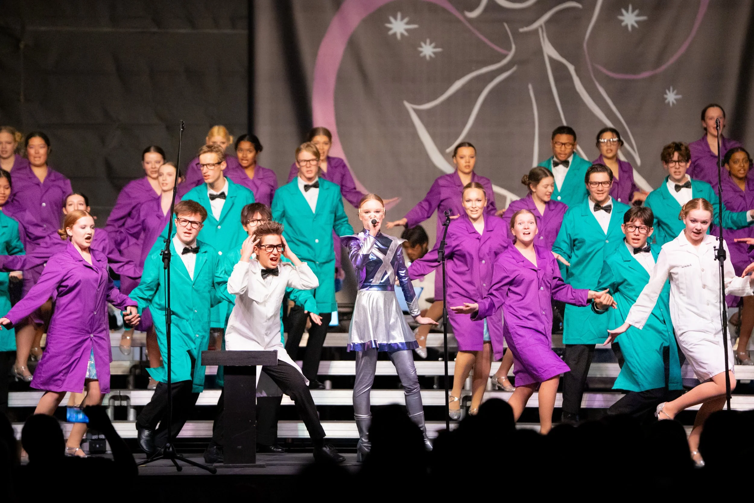A group of children performing on stage, dressed in colorful costumes, with a girl in metallic silver standing at the front holding a microphone, and a backdrop with abstract star and moon designs.