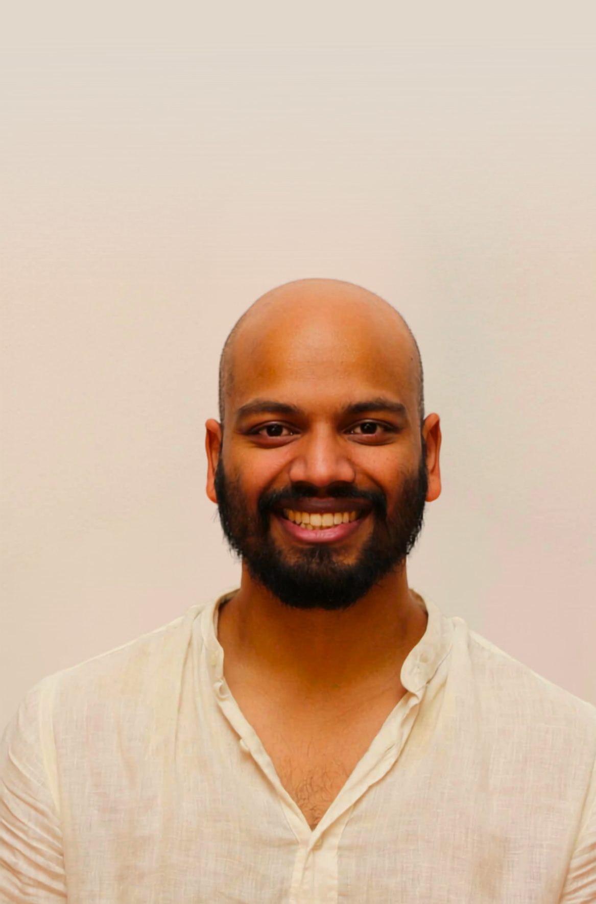 Smiling man with a beard and a shaved head wearing a light-colored shirt against a plain background.