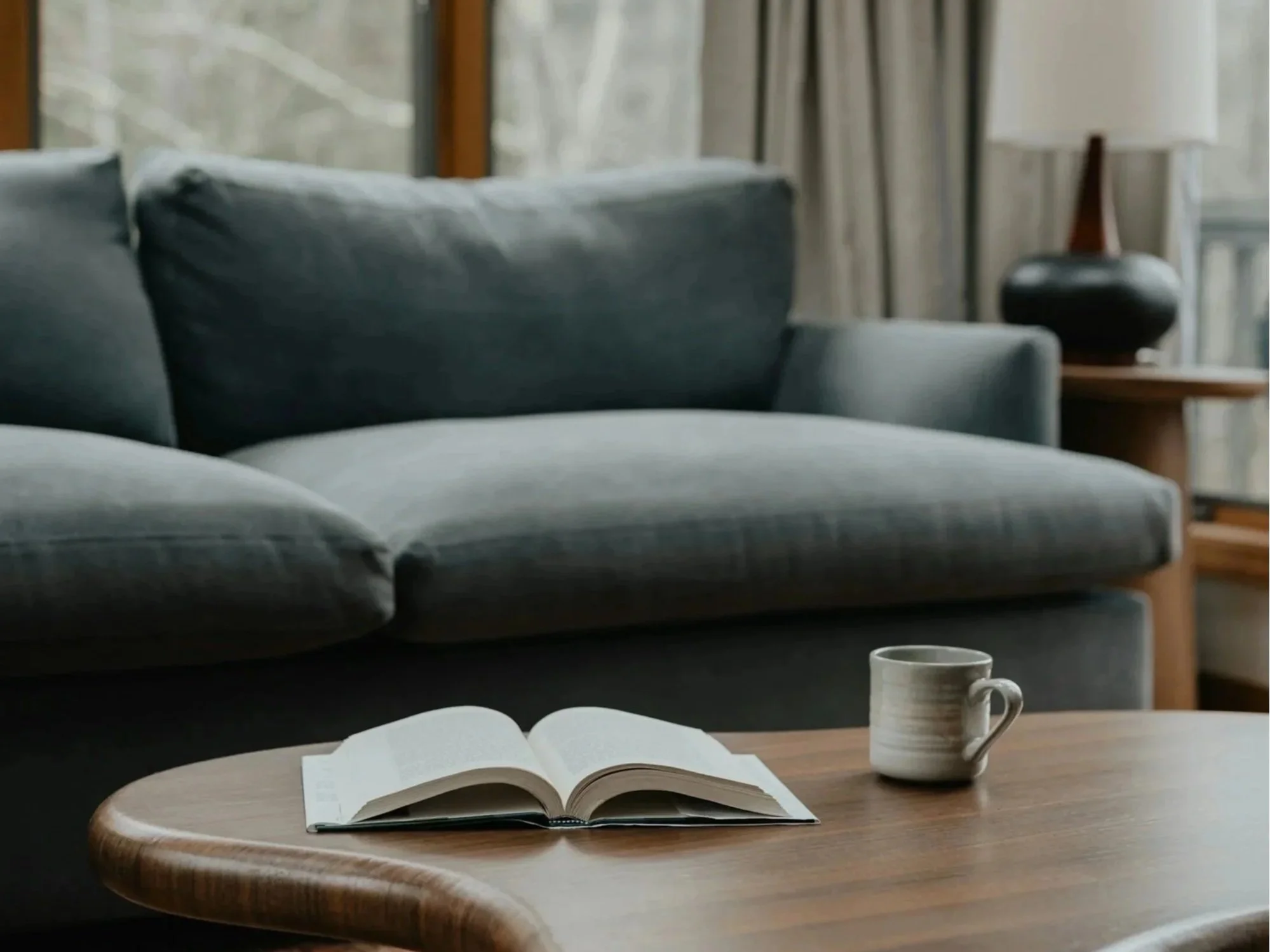 Counselling room scene with a couch, an open book on a wooden coffee table, a mug, and a side table with a lamp, large windows with curtains, and a decorative vase.