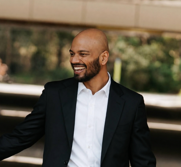 A warm and welcoming smiling man in a black suit and white shirt walking outdoors.