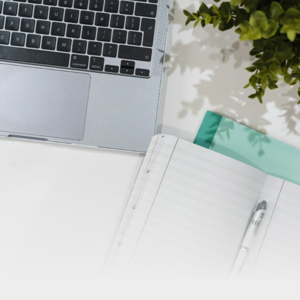 Open laptop keyboard, open notebook with lined pages and pen, green plant in top right corner, on white desk.