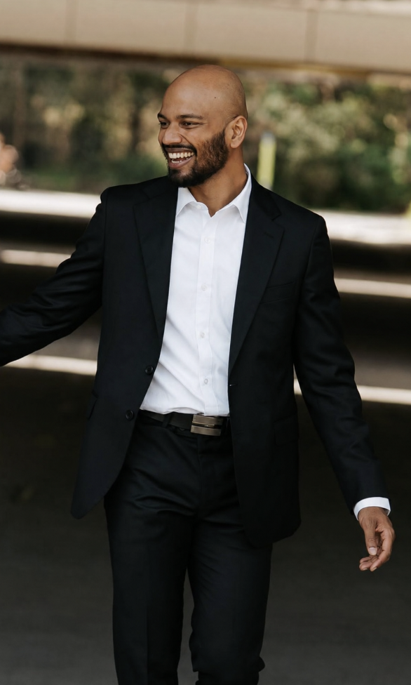 A warm and welcoming smiling man in a black suit and white shirt walking outdoors.