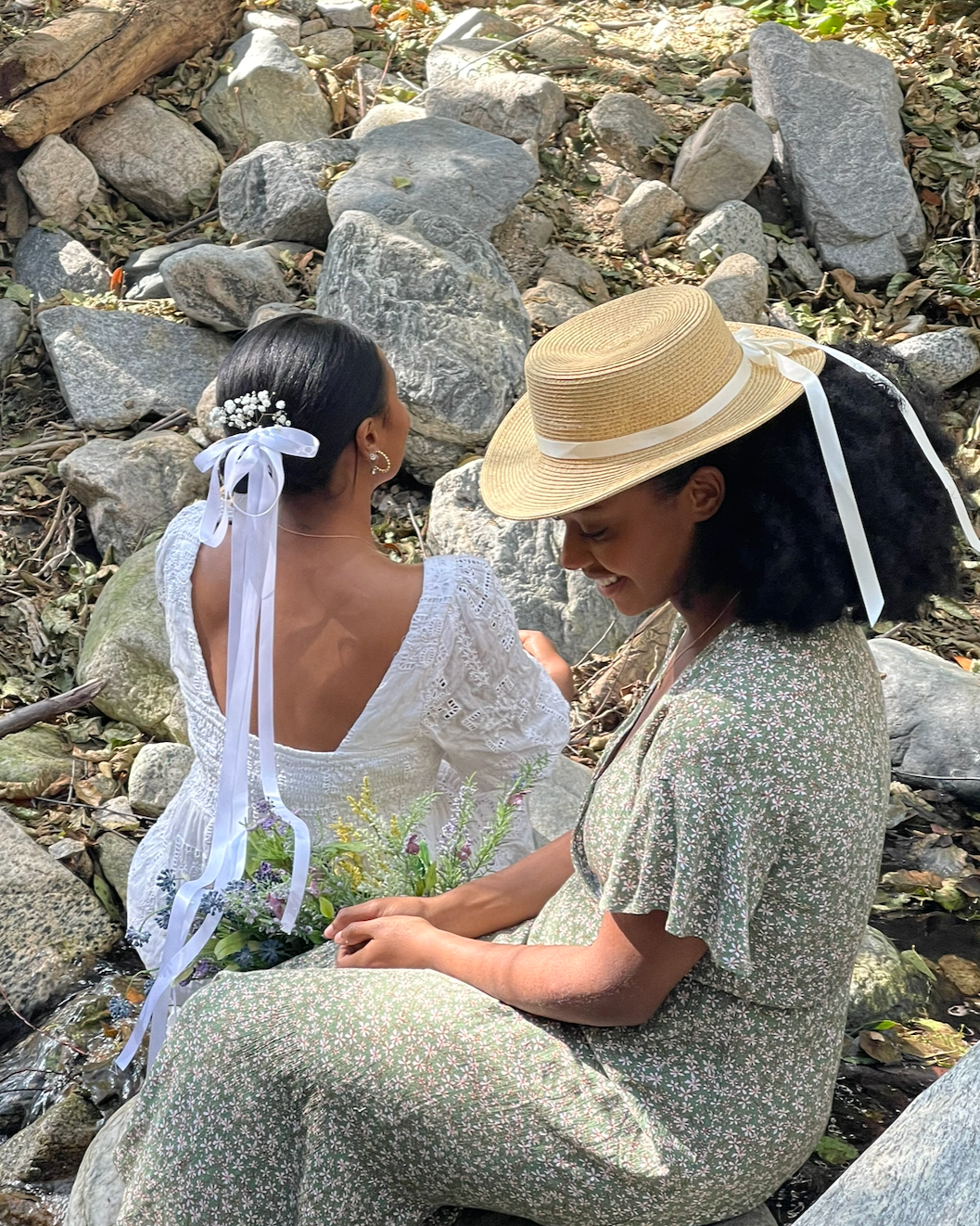 Two women sitting on rocks in a natural outdoor setting, one wearing a straw hat and the other with flowers in her hair. They are dressed in light-colored, summery clothing and holding a bouquet of flowers, engaging in a joyful moment.