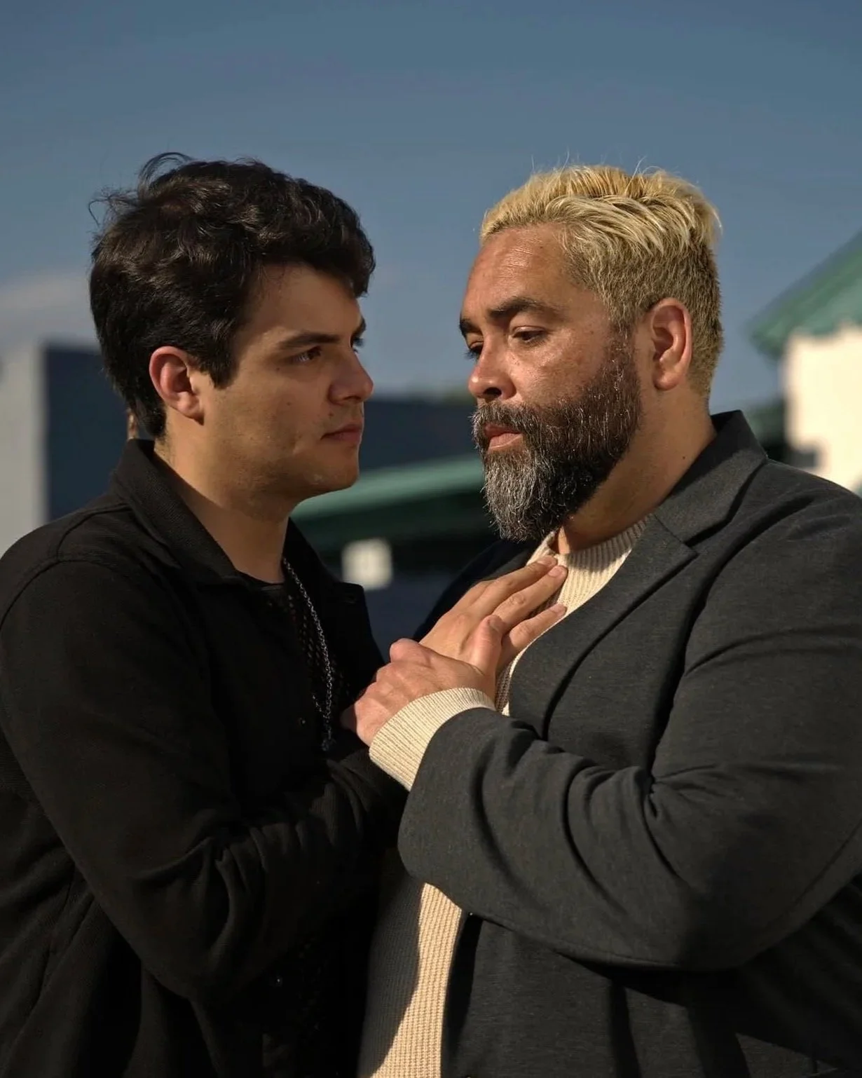 Two men face each other closely, one with dark hair and the other with blonde hair and a beard, holding each other's shoulders and gazing intensely.