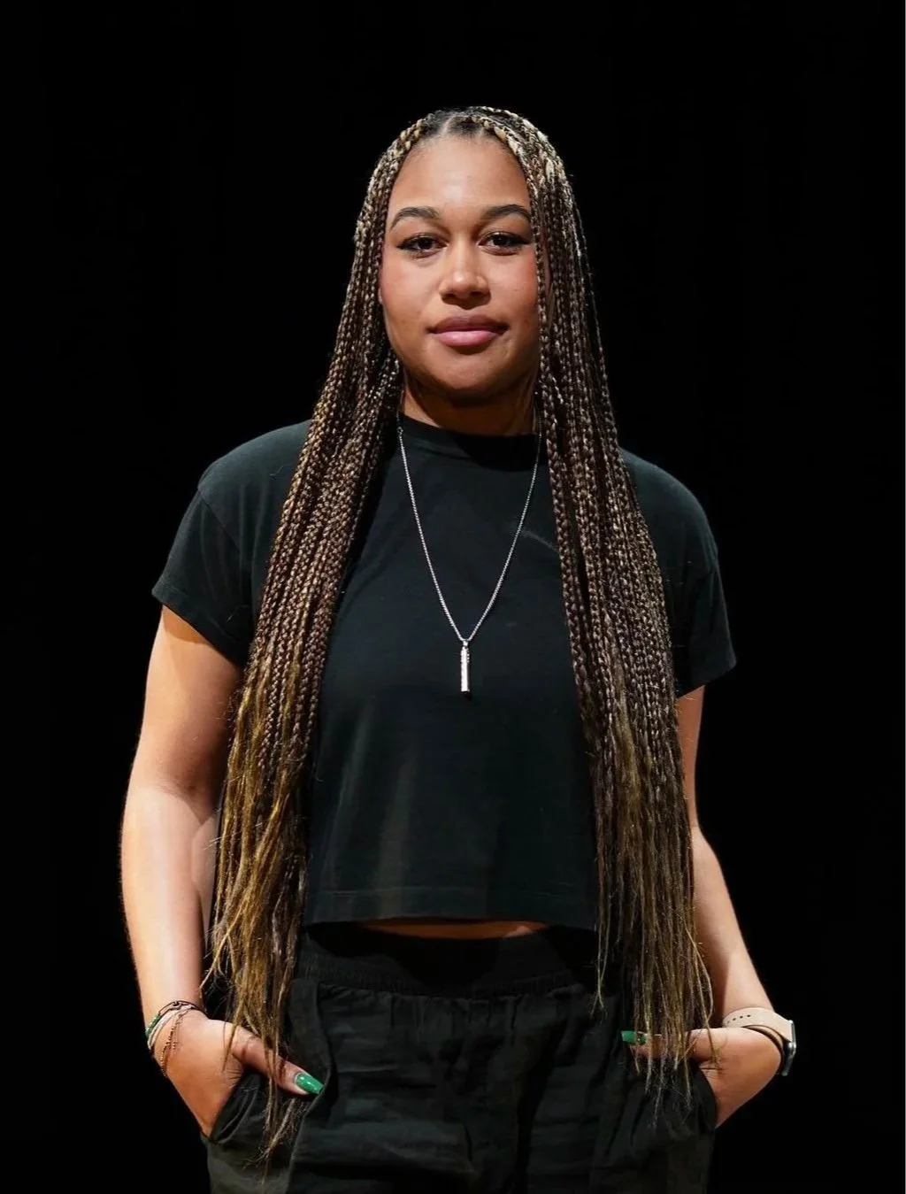 A young woman with long braids wearing a black t-shirt, black pants, and a necklace, standing with her hands in her pockets against a black background.