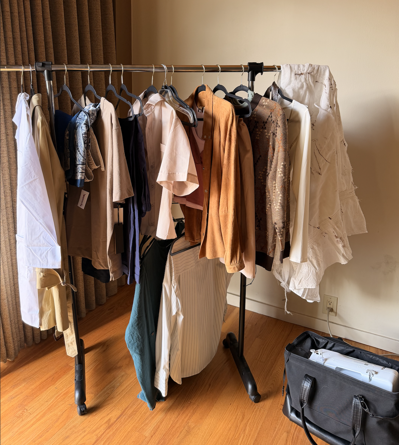 Clothes hanging on a portable clothing rack, including shirts, jackets, pants, and skirts, with some on hangers and a black duffle bag on the floor nearby.