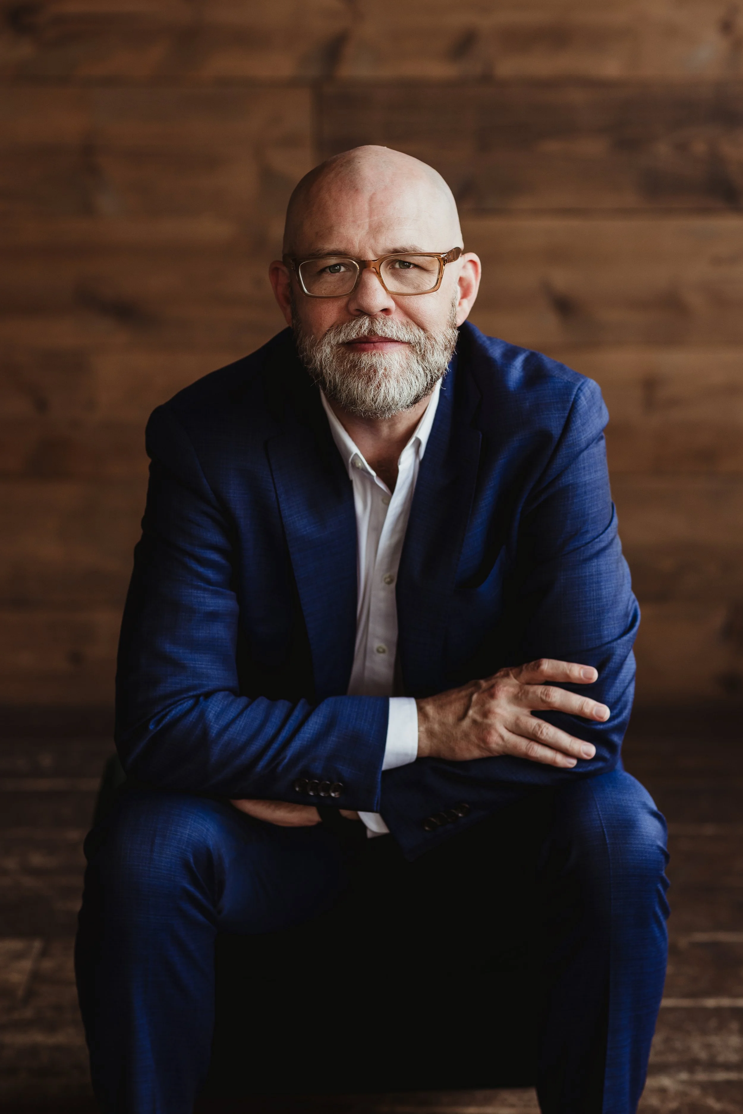 A middle-aged man with a beard, glasses, and a bald head, wearing a blue suit with a white shirt, sitting against a wooden background.