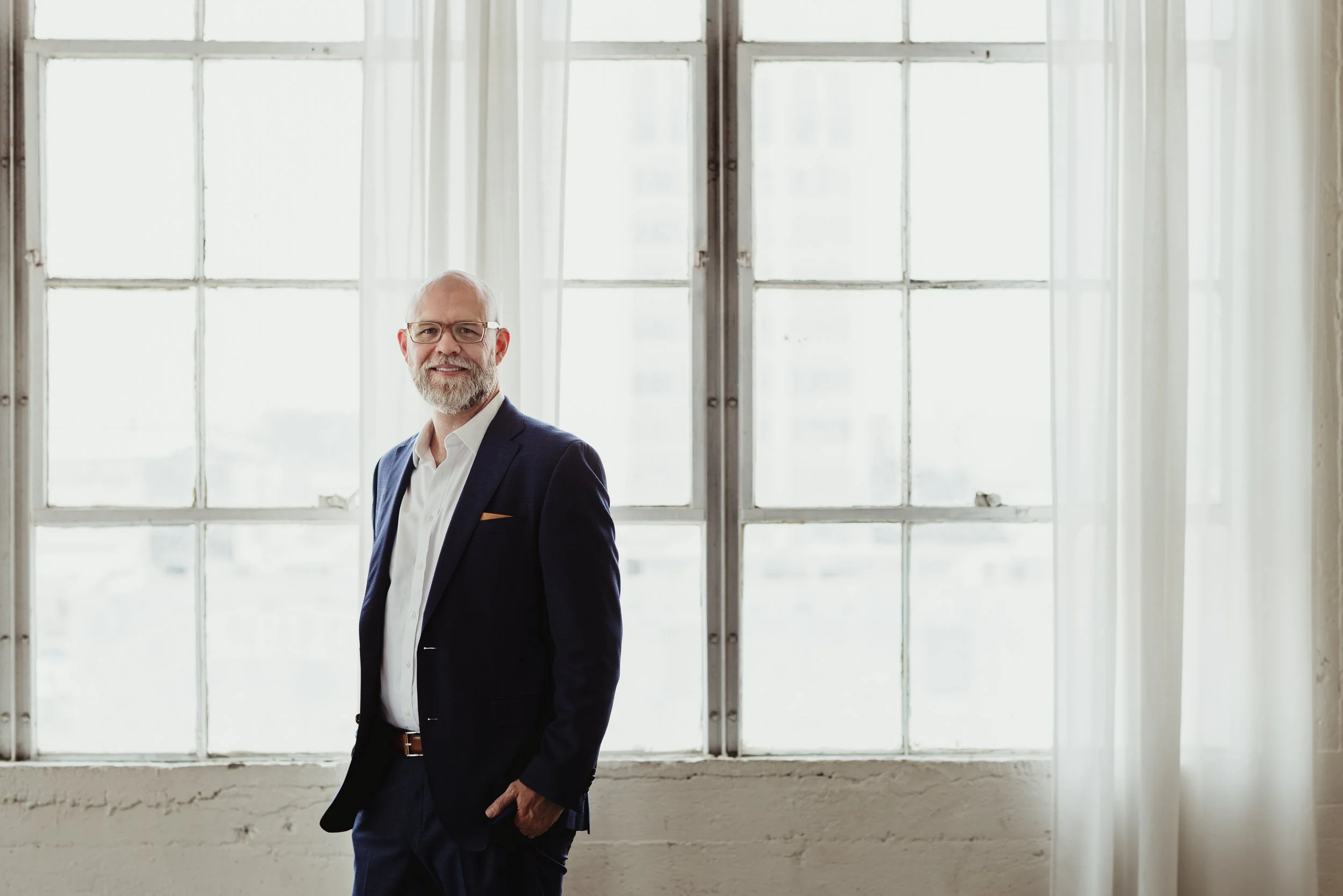 A middle-aged man with glasses and a beard, dressed in a dark suit and white shirt, standing in front of large windows with sheer curtains in a bright room who is a car industry expert.