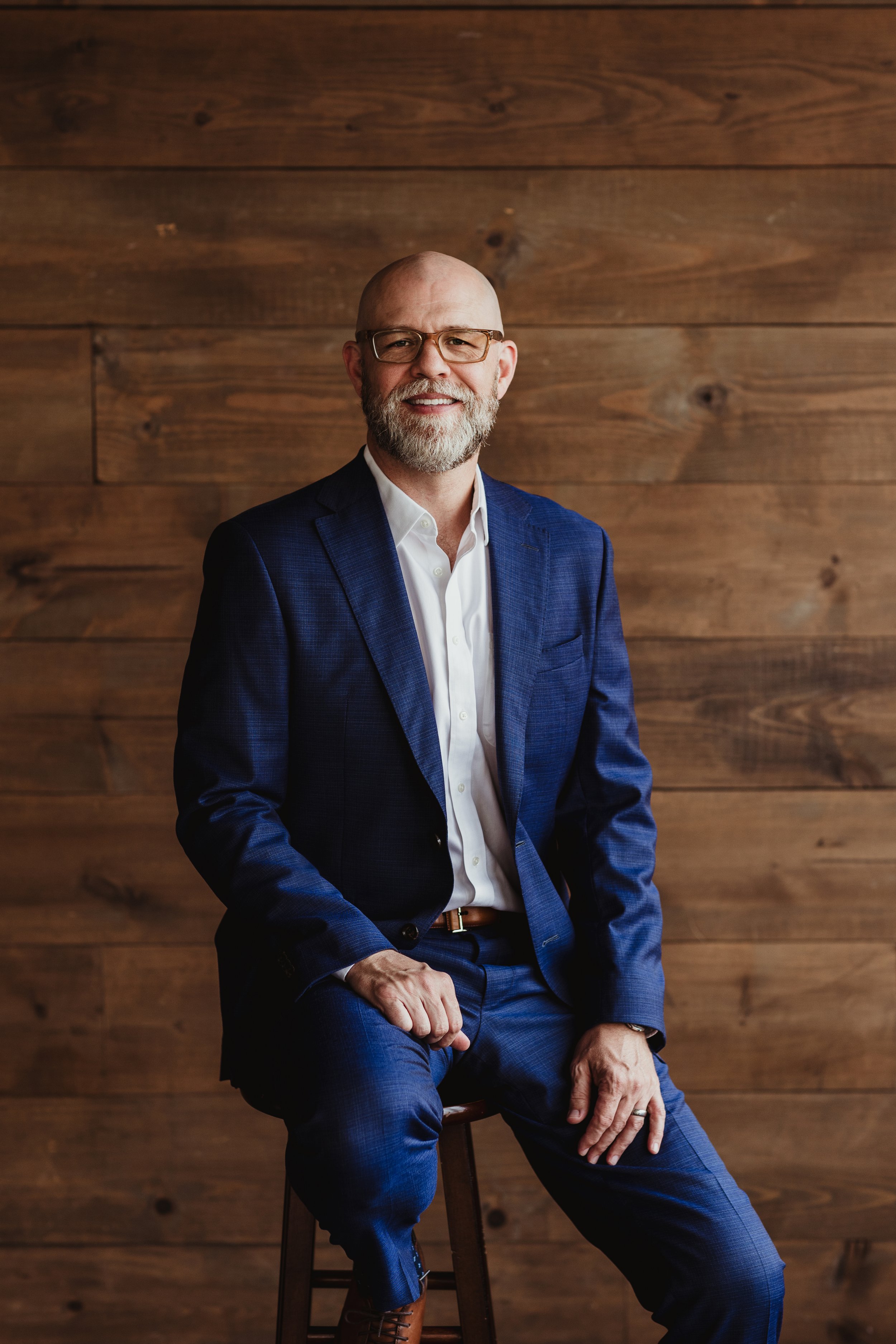 A middle-aged man with a beard and glasses, wearing a navy blue suit and white shirt, sitting on a wooden stool against a wooden wall.