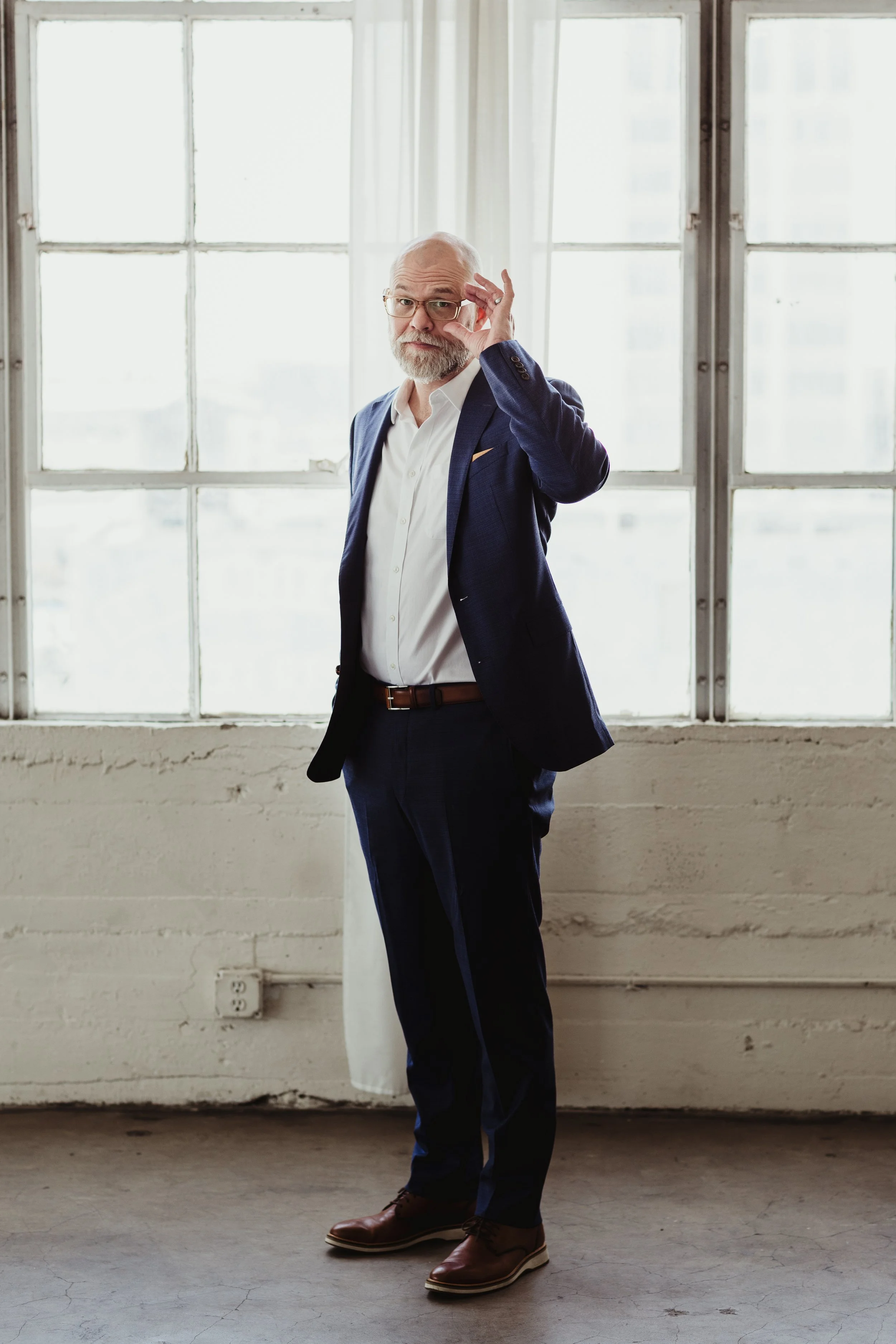 A middle-aged man with a grey beard and bald head, wearing glasses, a white shirt, a dark blue suit jacket, and brown shoes, is standing in front of a large window with white curtains in a room with a concrete floor and exposed brick wall. He is adjusting his glasses with his right hand.