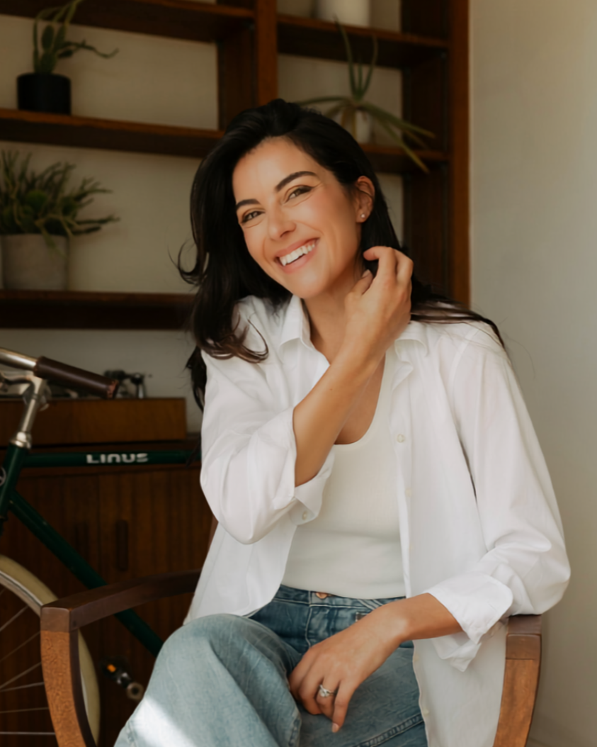 A woman with dark hair and a white shirt smiling while sitting indoors in front of a wooden shelf with potted plants.