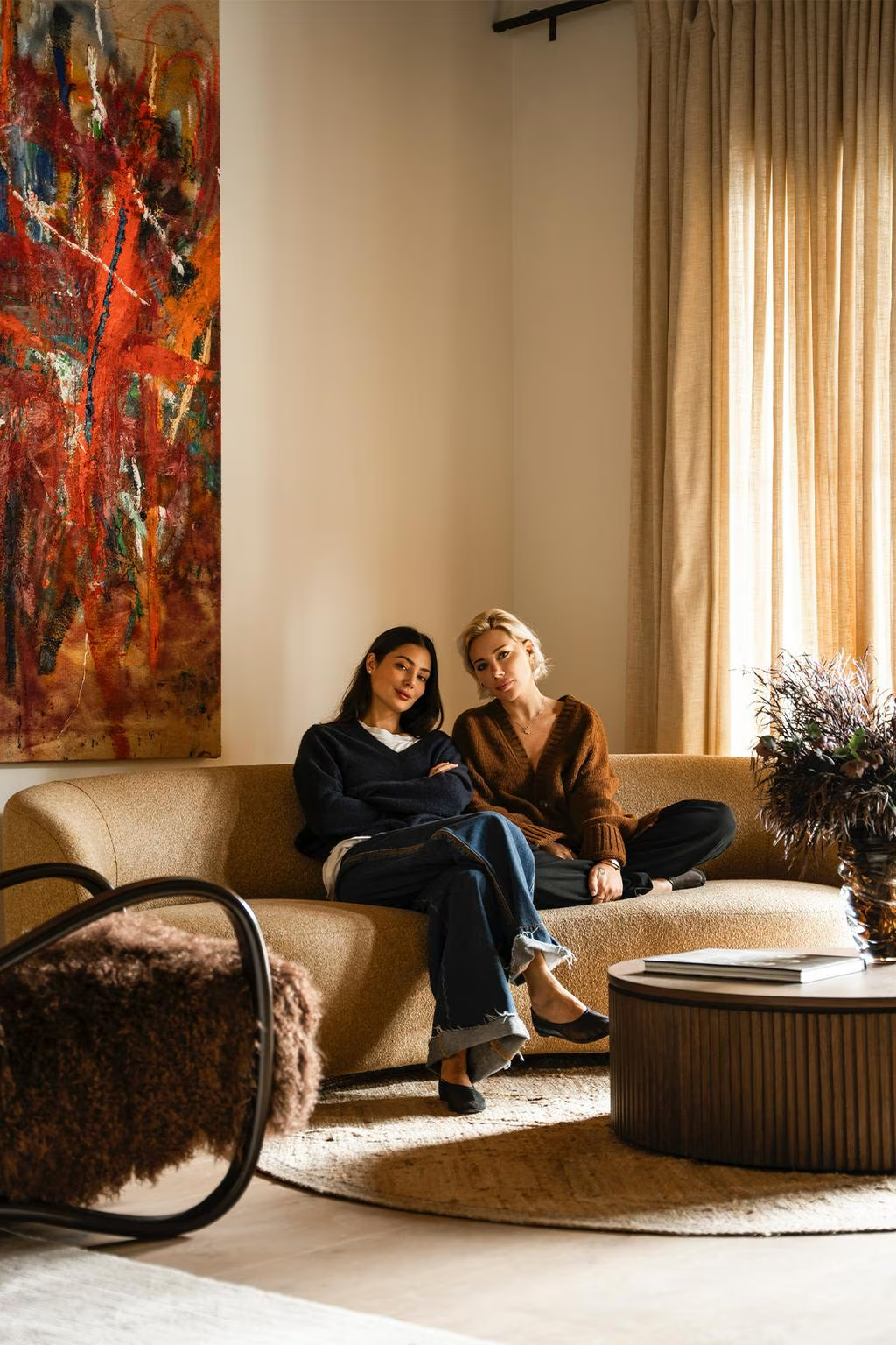 Two women sitting on a beige sofa in a living room with abstract wall art and a large window with beige curtains.