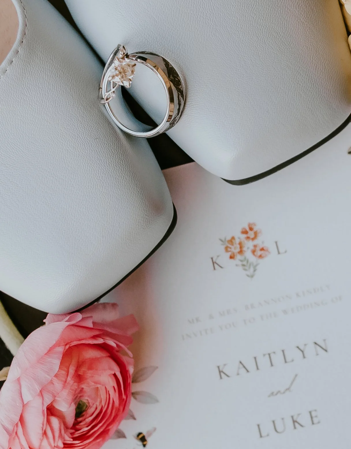 Two wedding rings sit on the toe of a light blue shoe. There is a pink flower next to the shoe and a wedding invitation for Kaitlyn and Luke's wedding. 