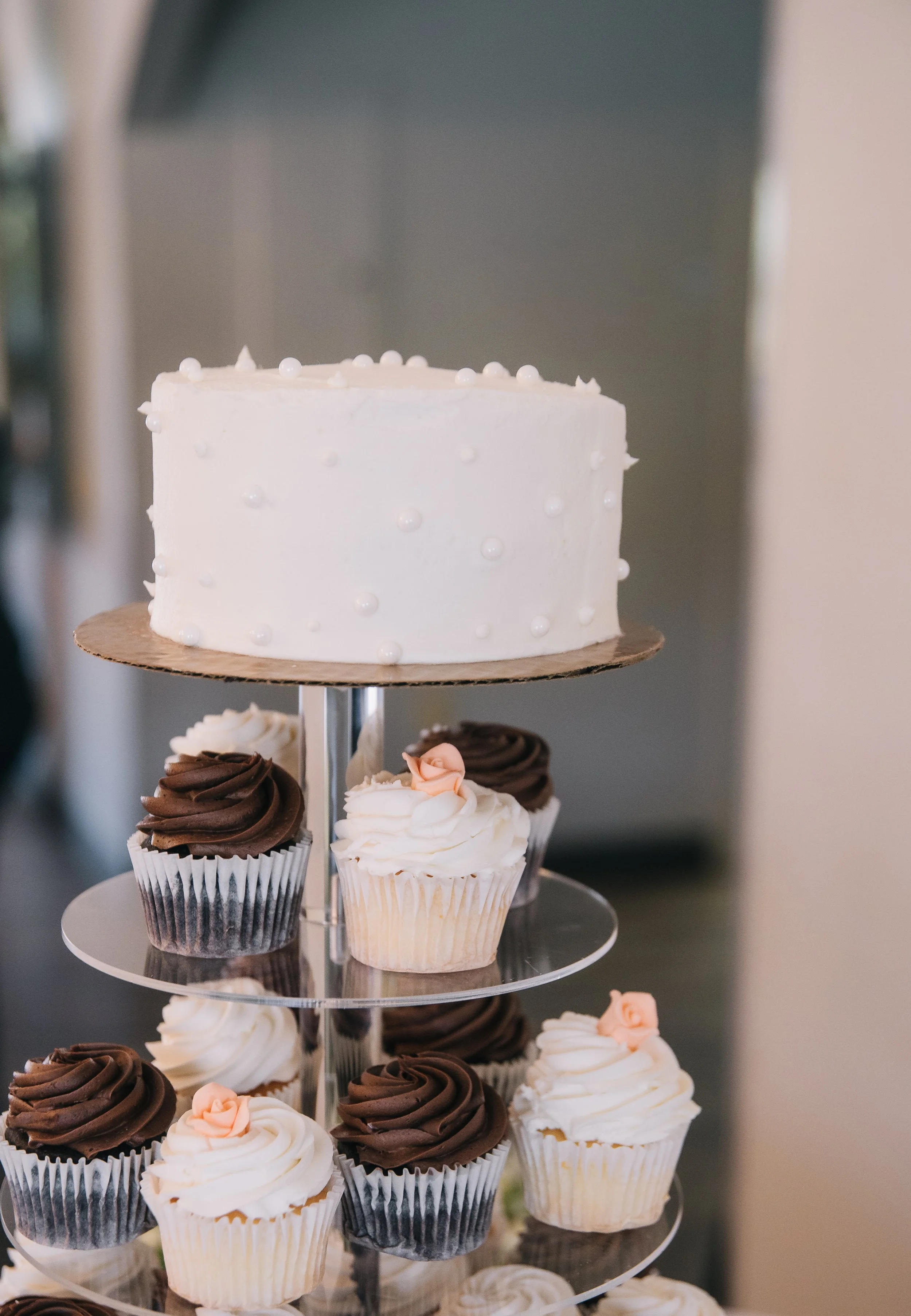 A white cake with small white edible pearls on a layered dessert stand, surrounded by chocolate and vanilla cupcakes with frosting and small decorative roses.