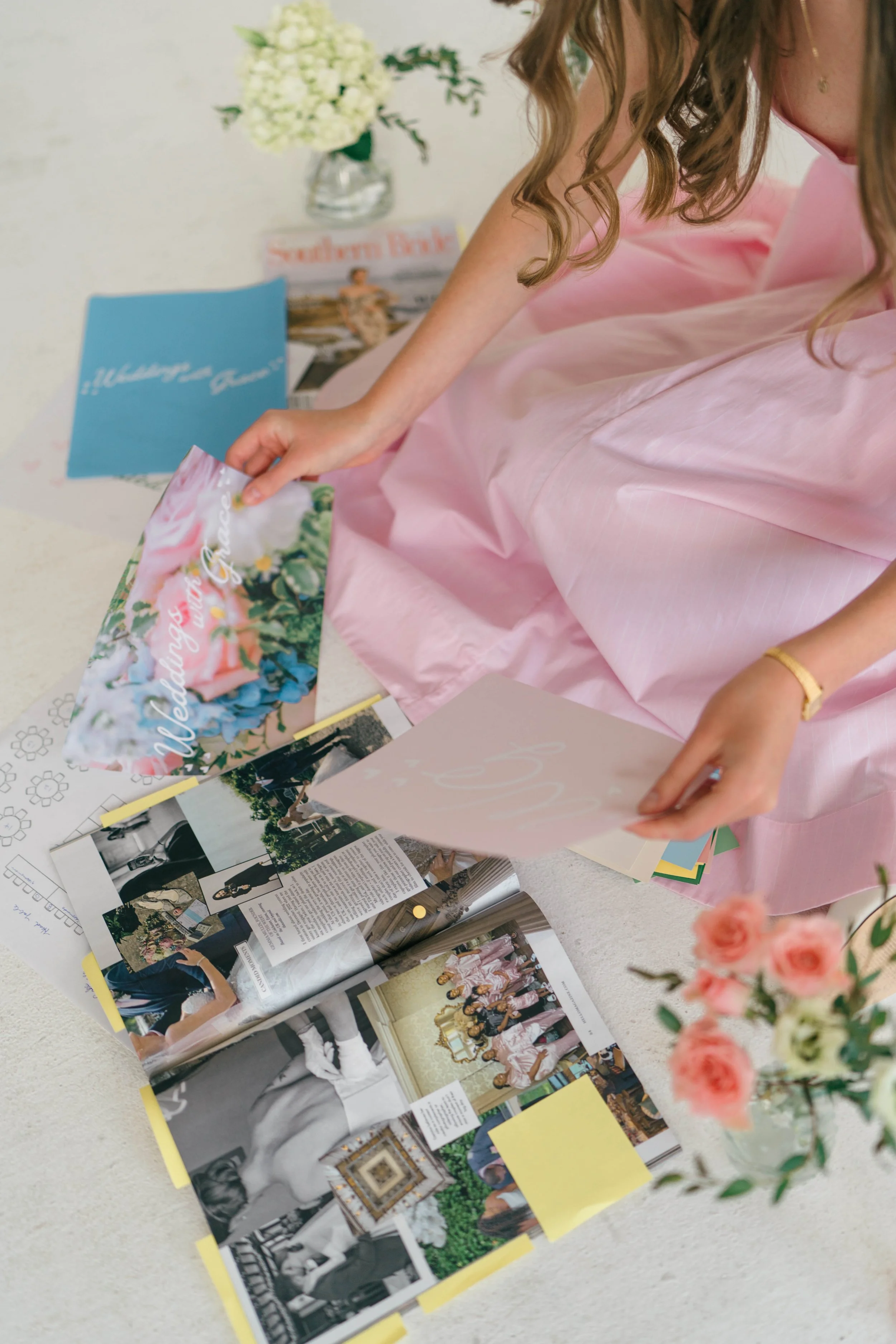 A woman in a pink dress is looking through wedding magazines and photos spread out on a table. There are flowers and other decorative items in the background.