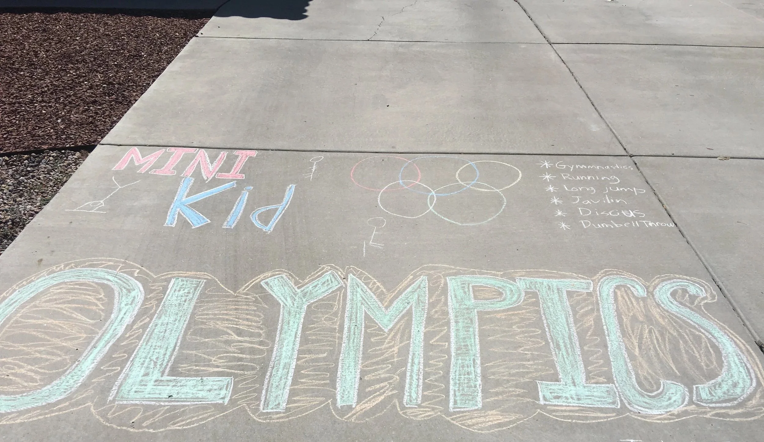 Chalk drawing on concrete sidewalk with the words 'Mini Kid Olympics,' alongside a drawing of a figure with a kite, a list of activities including gymnastics, running, long jump, discus, and javelin throw.