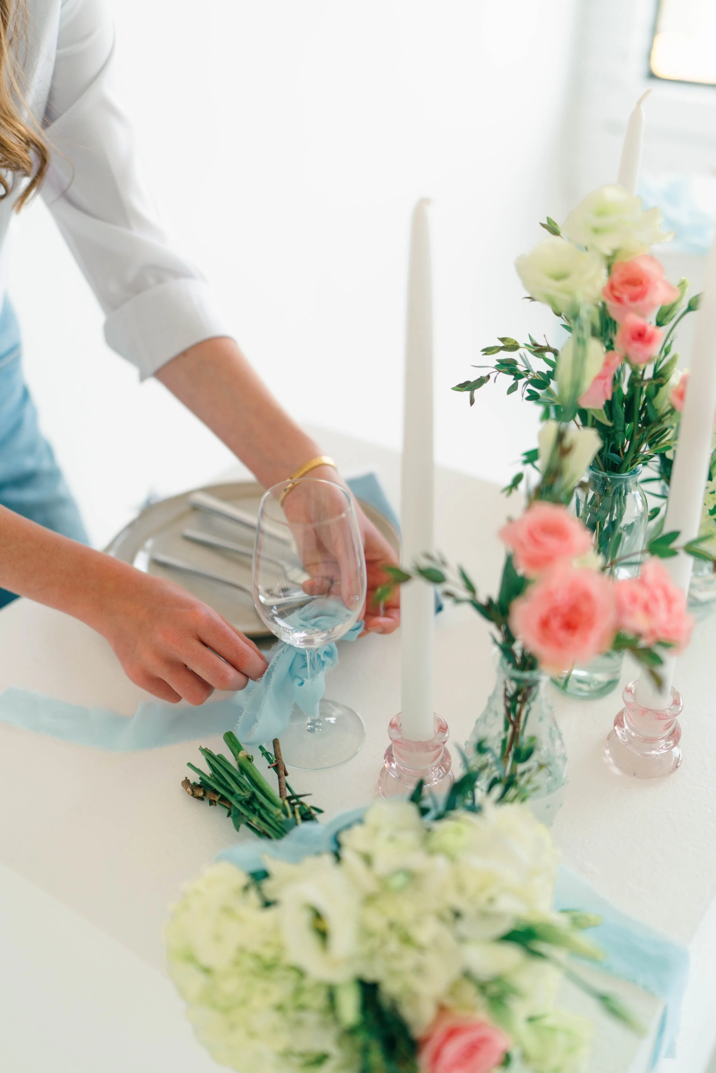 Grace Veazey preparing floral centerpieces and candles, arranging a glass with a blue ribbon on a white table.