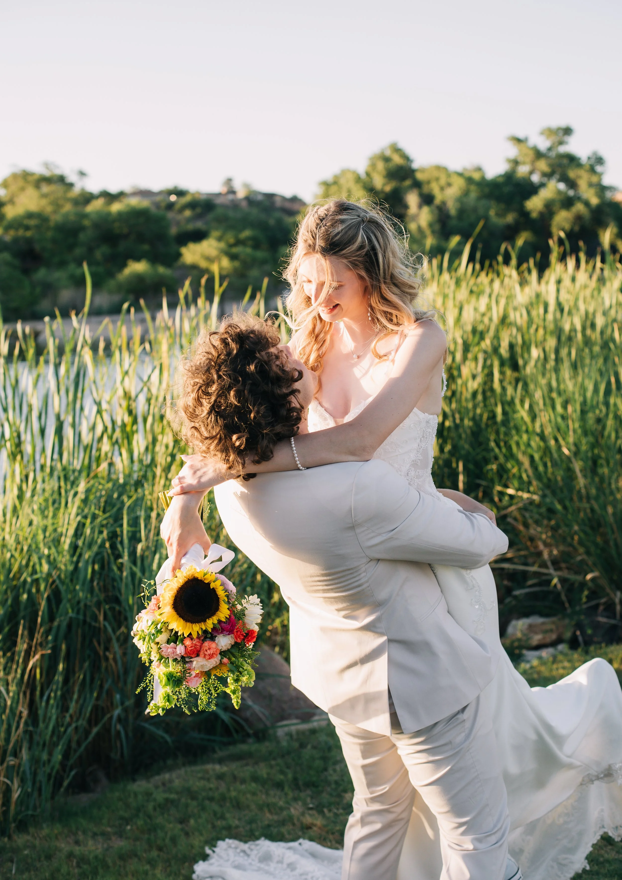 A bride in a wedding dress holding a bouquet of flowers, being lifted by a groom in a tan suit outdoors during sunset, with green trees, a hill, and a lake in the background.