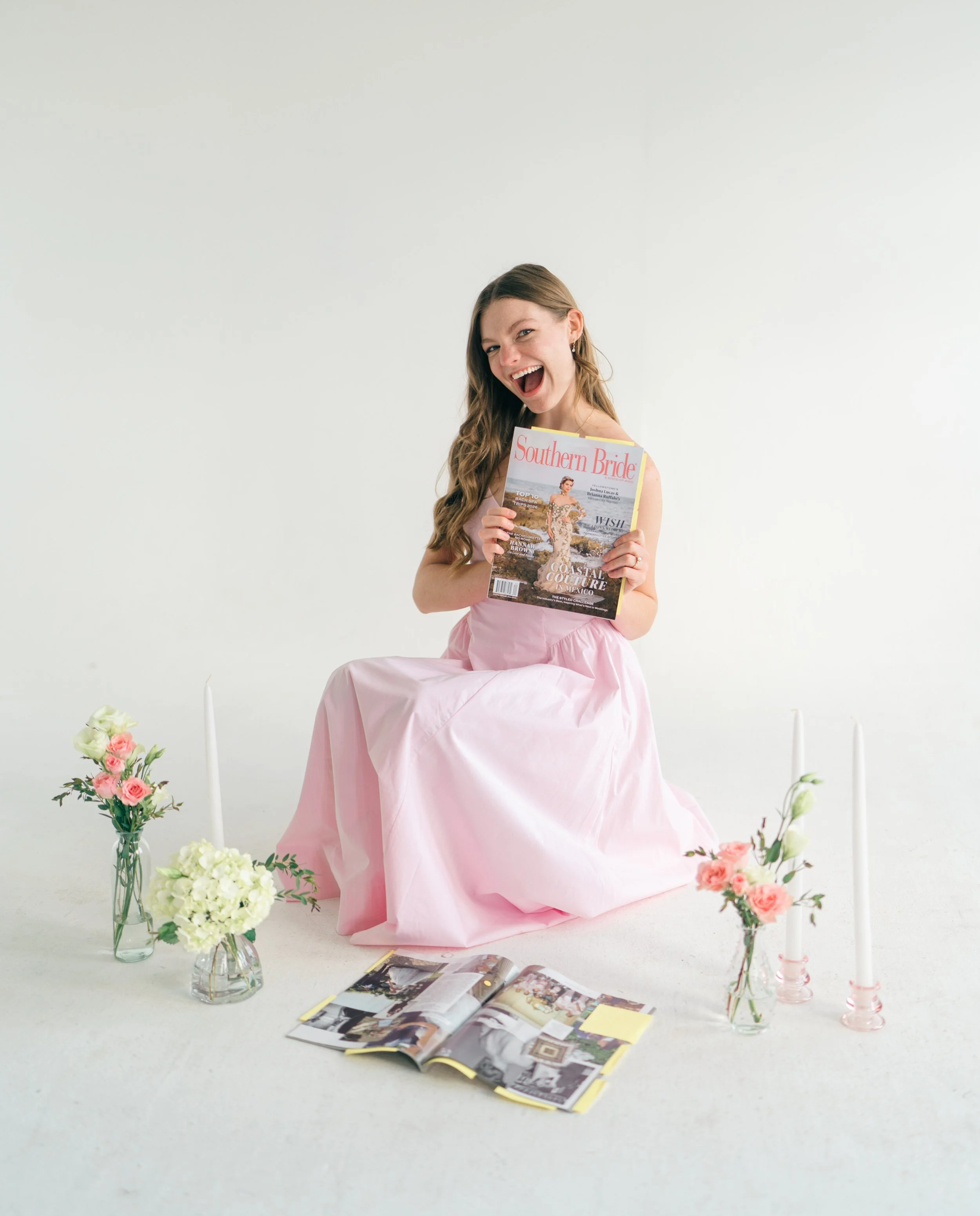A young woman with long hair, sitting on the floor, holding a magazine titled 'Southern Bride', and smiling at the camera. She is surrounded by pink and white flowers in vases, candles, and magazines, with a plain white background.