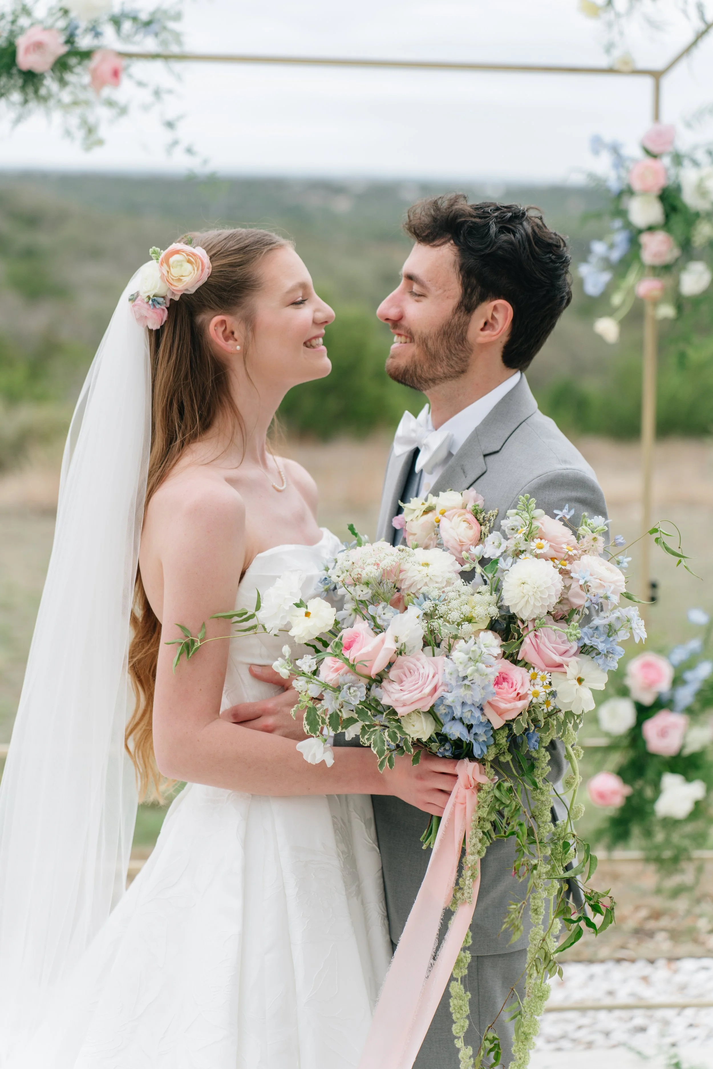 A bride and groom smiling and looking at each other during their wedding ceremony outdoors, with a floral arch and scenic landscape in the background.