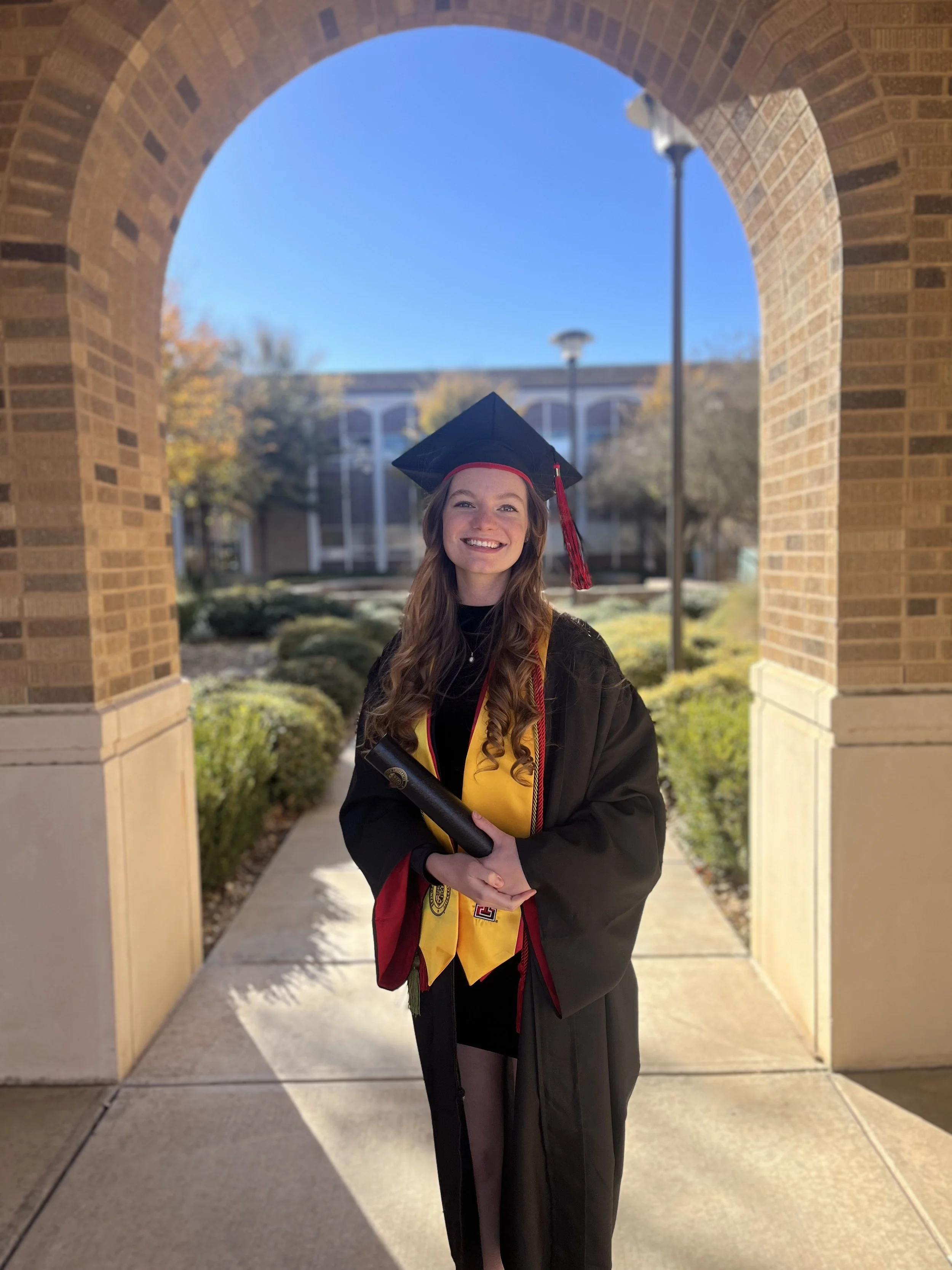 Grace Veazey in graduation attire smiling and holding a diploma under an archway at Texas Tech University.
