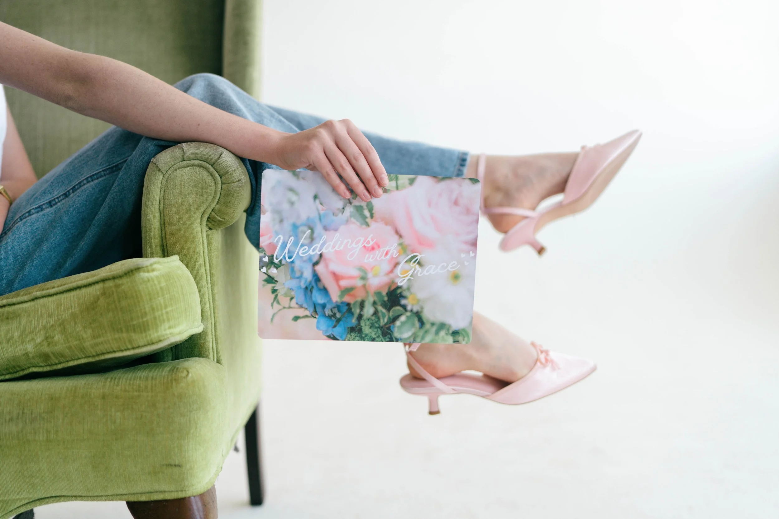 Close-up of a Grace Veazey sitting on a green velvet chair, holding a sign with a floral background and the words "Weddings with Grace." She is wearing pink high-heeled shoes and light blue jeans.