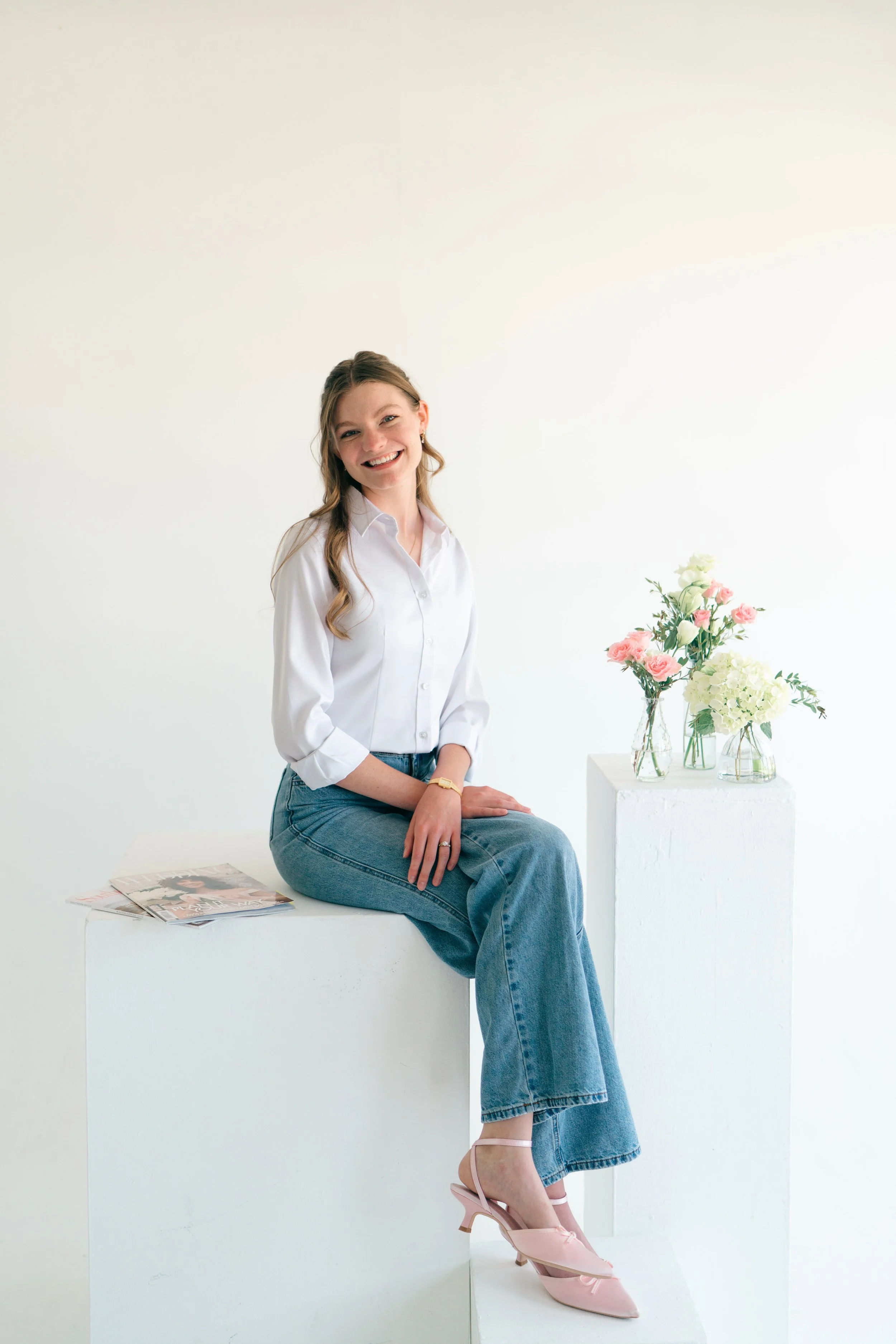 Grace Veazey sitting on a white block with a white background. She is wearing blue jeans, a white shirt, and pink heeled shoes. There are floral vases and wedding magazines on the blocks next to her.