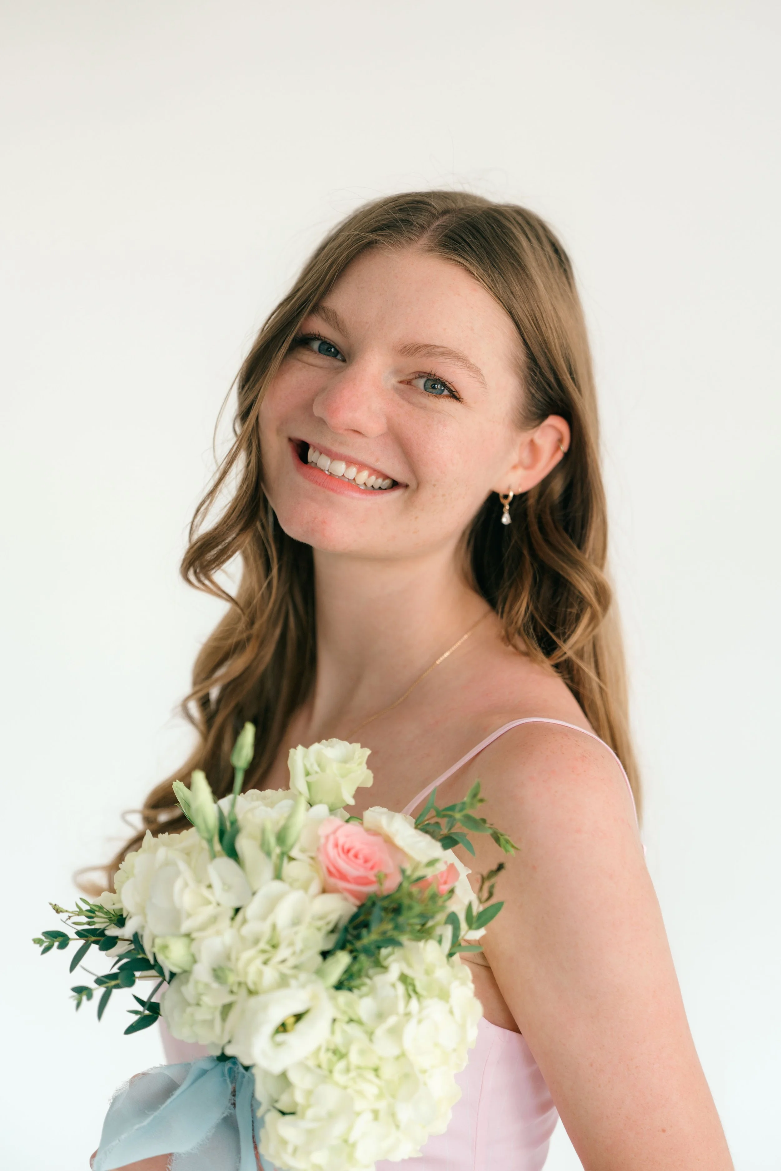 Grace Veazey, a young woman with wavy brown hair, blue eyes, and earrings, smiling while holding a bouquet of white and pink flowers against a plain white background.
