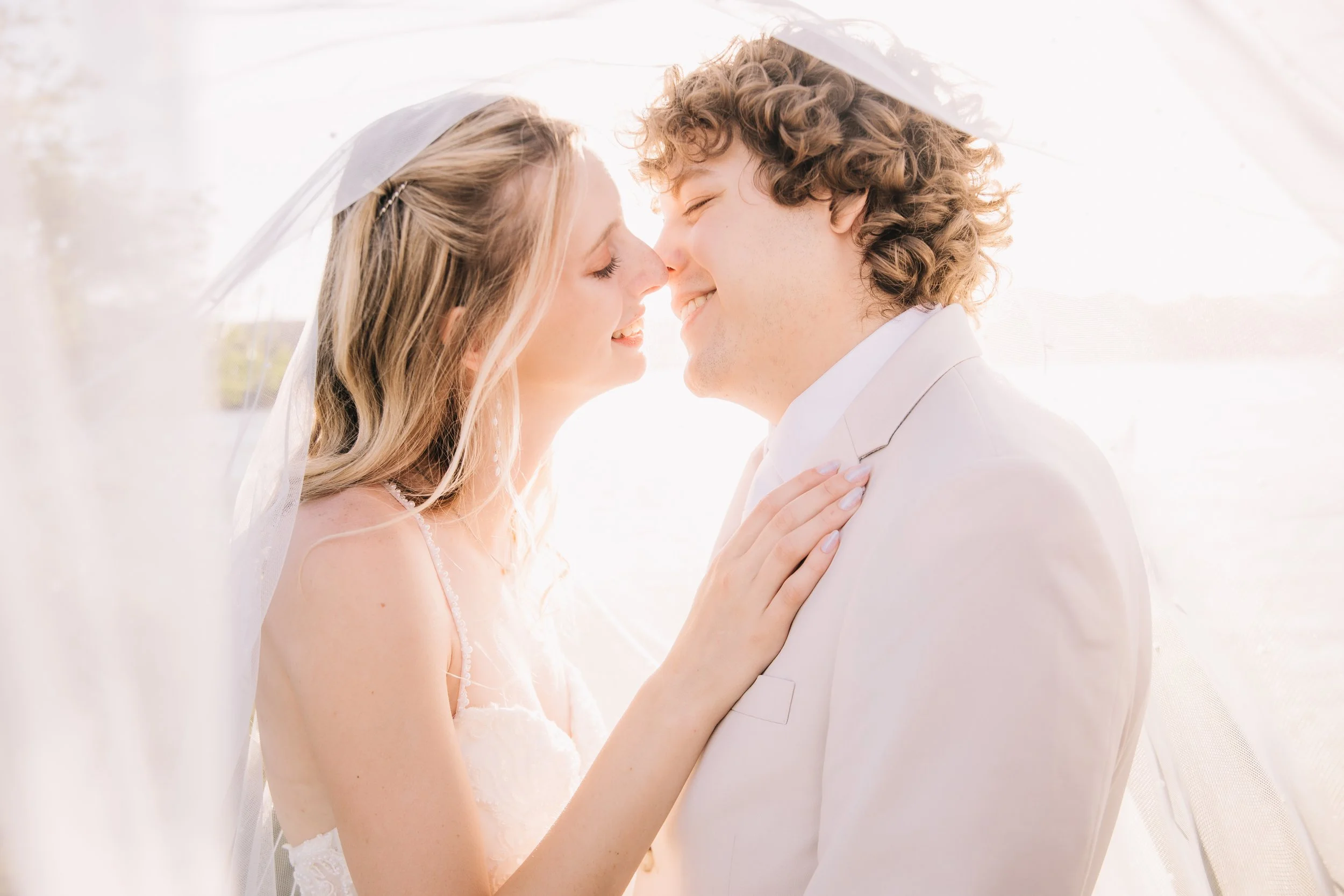A bride and groom are smiling with noses touching and eyes closed, standing under the bride's veil on their wedding day.