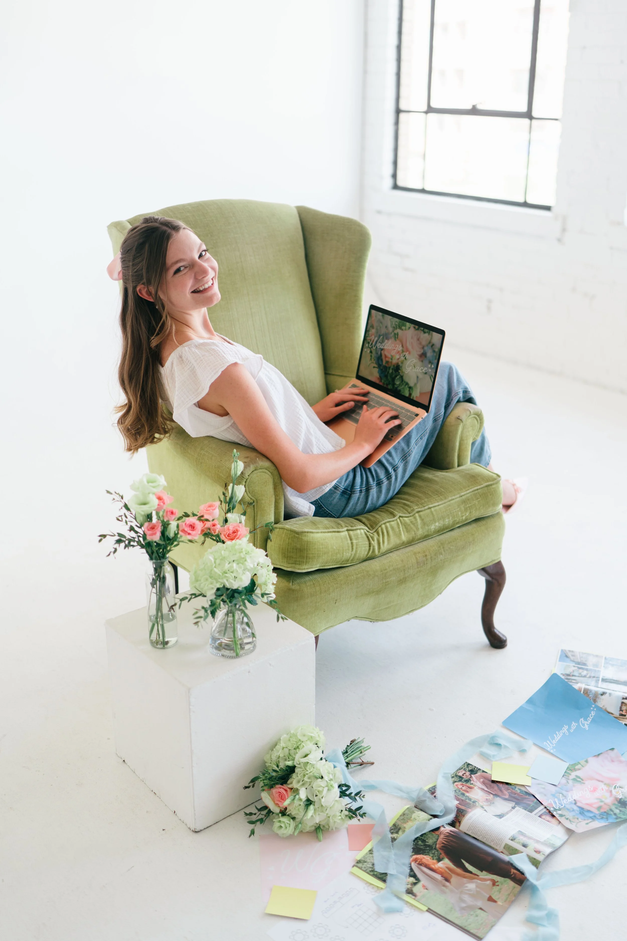 Grace Veazey, a woman with long brown hair sits on a light green armchair in a bright room, smiling, with flowers on a white table nearby and wedding planning materials scattered on the floor.