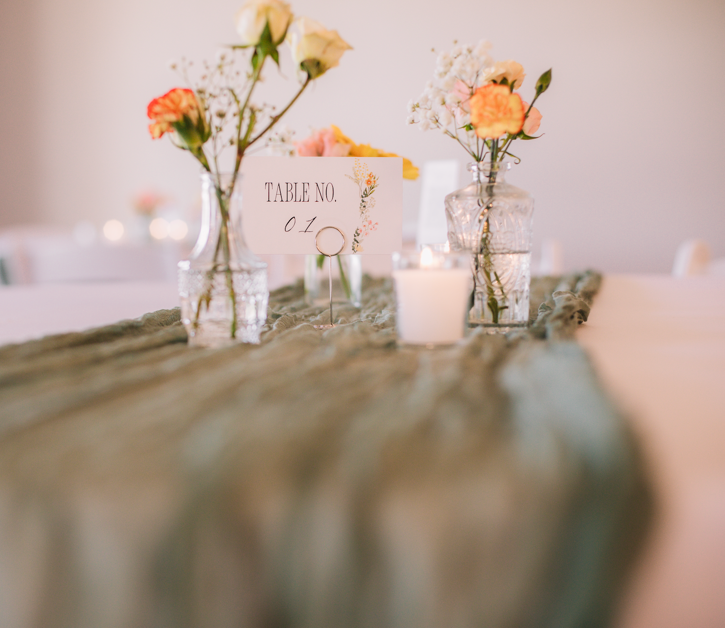 Wedding reception table centerpiece with glass vases holding white, pink, and orange flowers, a lit candle, and a table number sign.