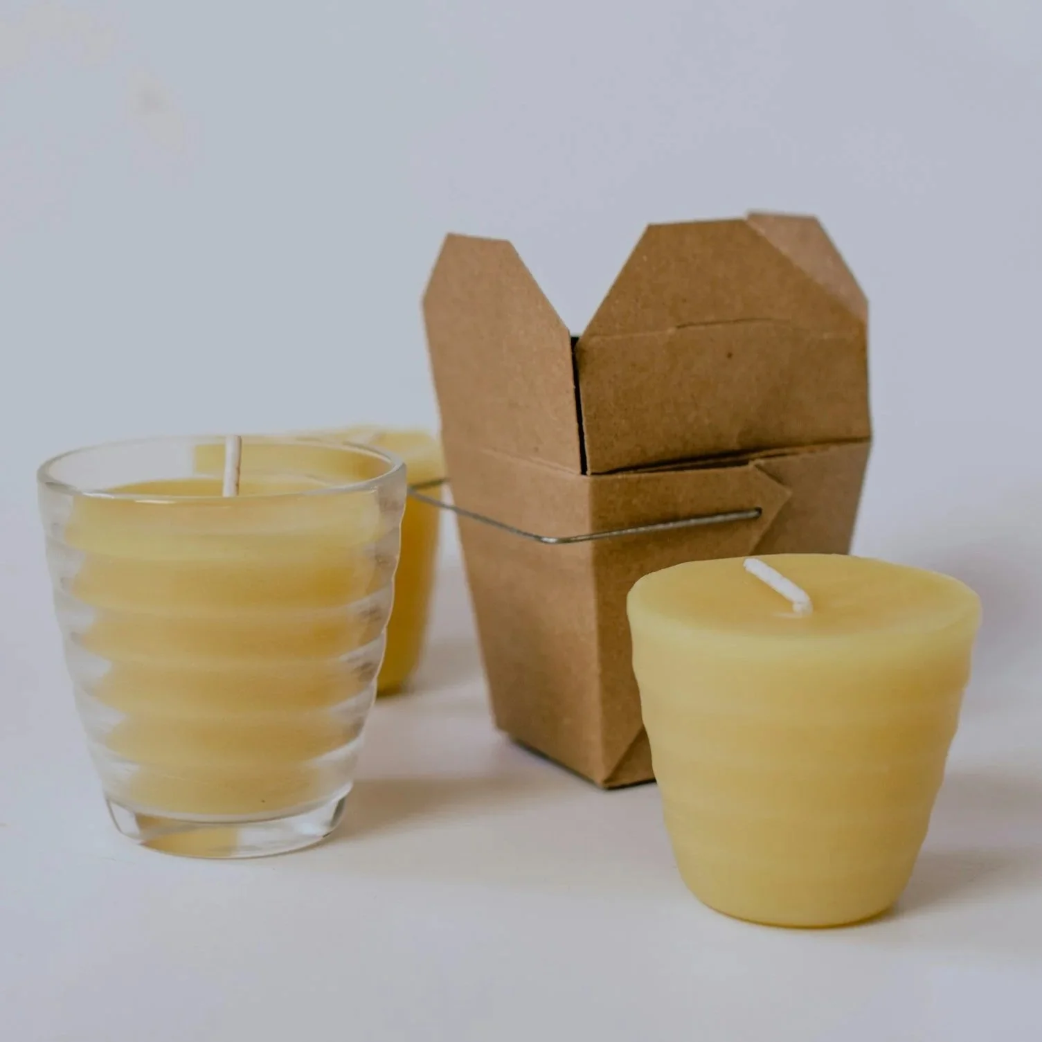 Two yellow candles and a glass candle holder next to a small cardboard box-shaped candle holder with a metal handle, all placed on a white surface.