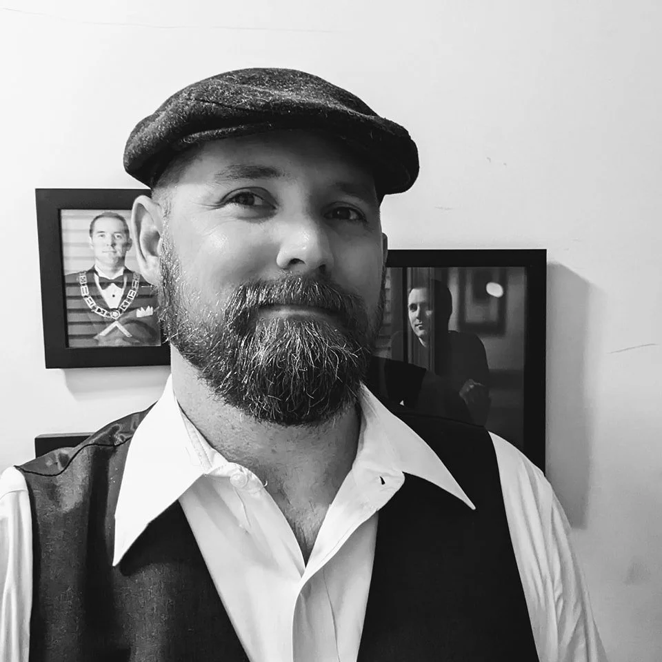 A man with a beard and mustache wearing a flat cap, white shirt, and vest, standing in front of framed photographs on a wall, with one photograph showing a woman.