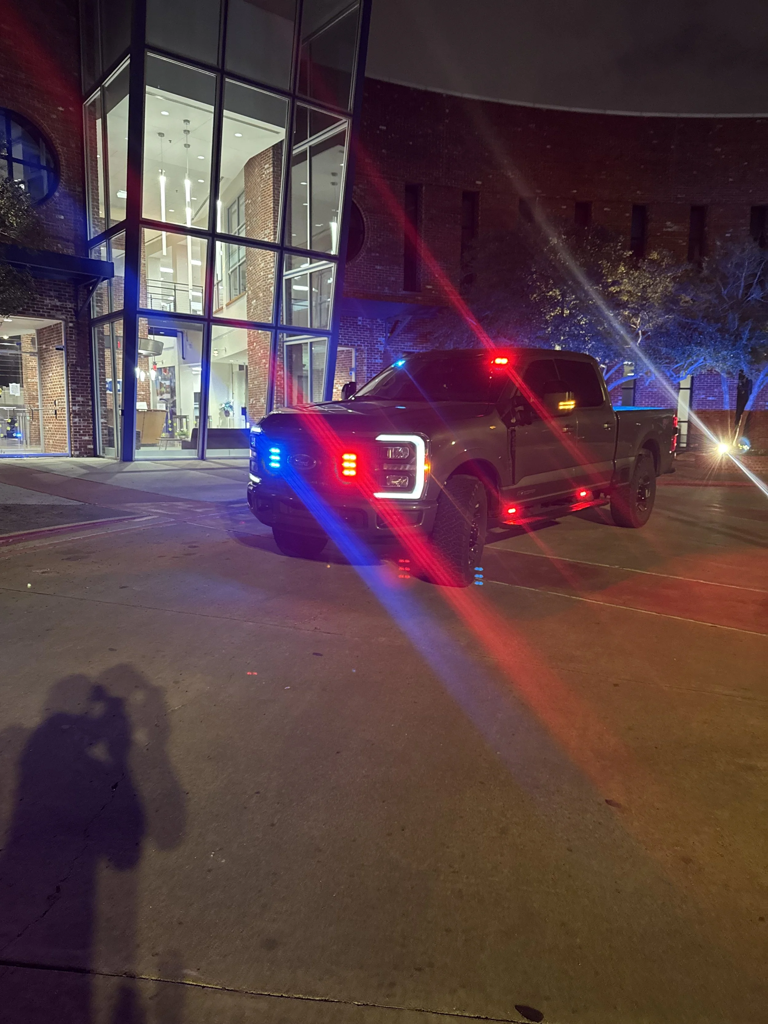 A dark-colored pickup truck with flashing red and blue lights is parked outside a brick building with large glass windows and a glass entrance. The photo is taken at night and the truck's lights are illuminating the area.