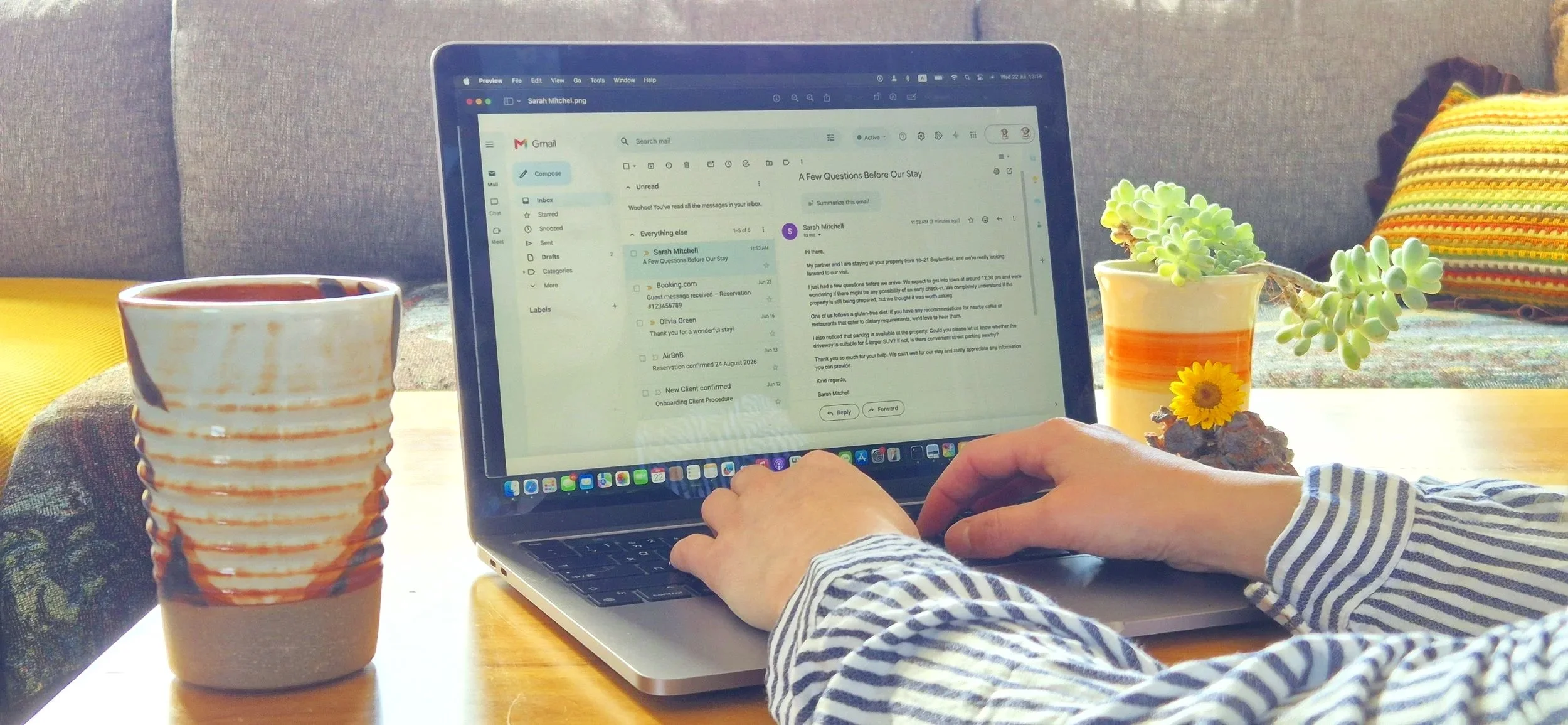 A person wearing a striped long sleeve shirt typing on a silver laptop with an email inbox open. The laptop is on a wooden table with a ceramic mug on the left and a small plant in a yellow pot on the right. There is also a colorful striped pillow and a grey couch in the background.