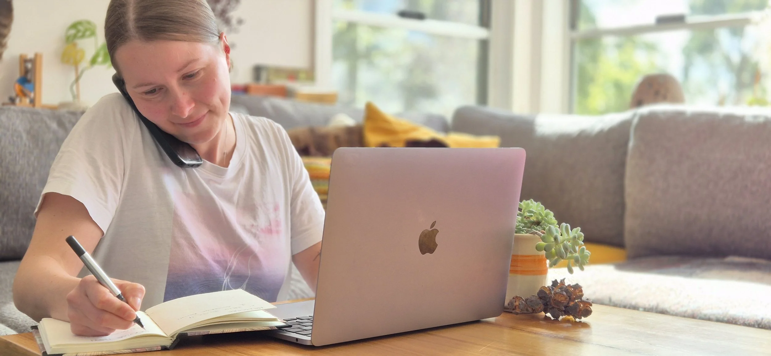 A woman with short hair, talking on a cellphone, writing in a notebook, at a desk with a laptop, in a cozy living room with large windows, a couch, plants, and a pile of crystals.
