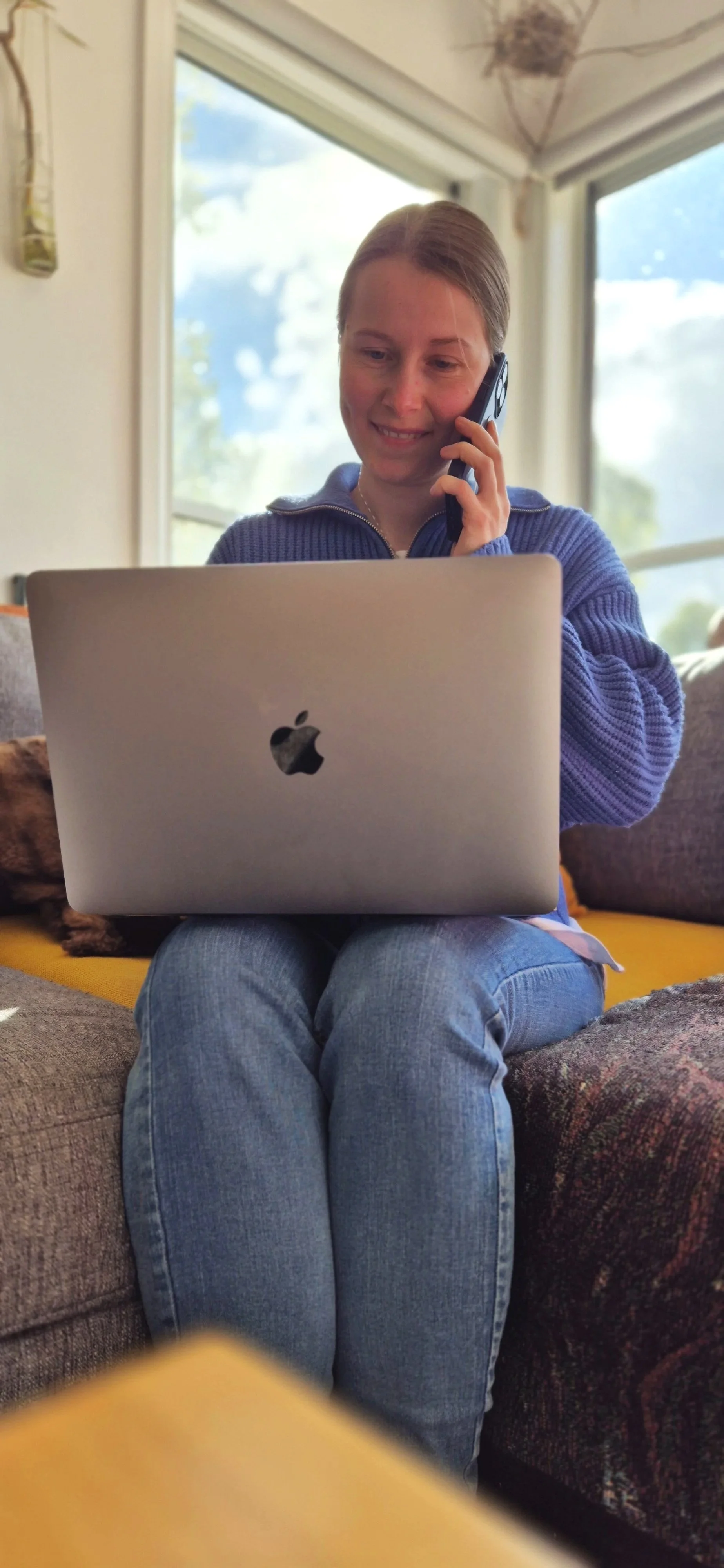 A woman sitting on a couch using a silver Apple MacBook and talking on a cellphone. There are large windows in the background with natural light coming in.