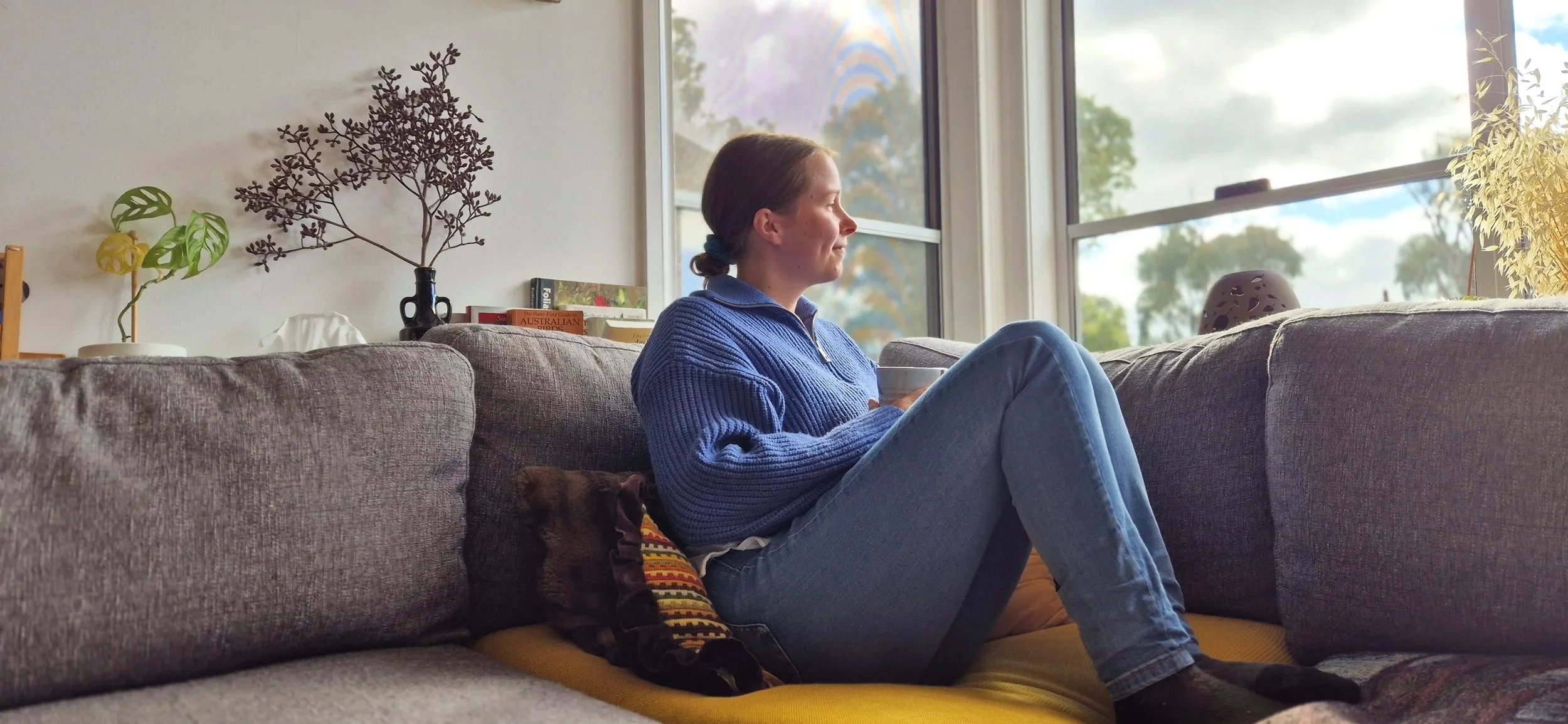 A young woman sitting on a gray couch in front of large windows, holding a mug, with a small dog resting beside her. Decor includes plants, books, and decorative items.