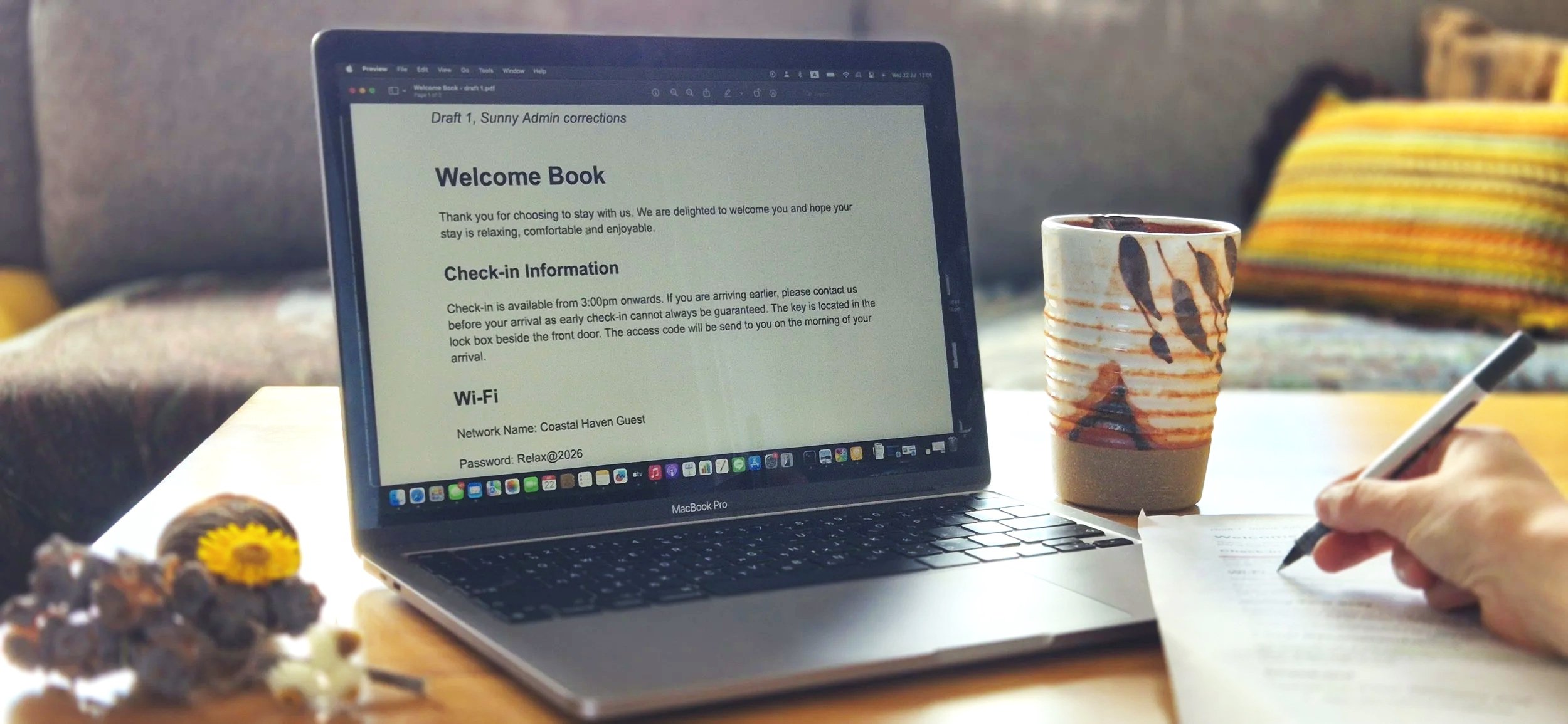 A laptop screen displaying a welcome book and check-in information, a person's hand writing on paper with a pen, a ceramic mug with a creative design, a colorful striped pillow on a couch in the background, and some food on the table.